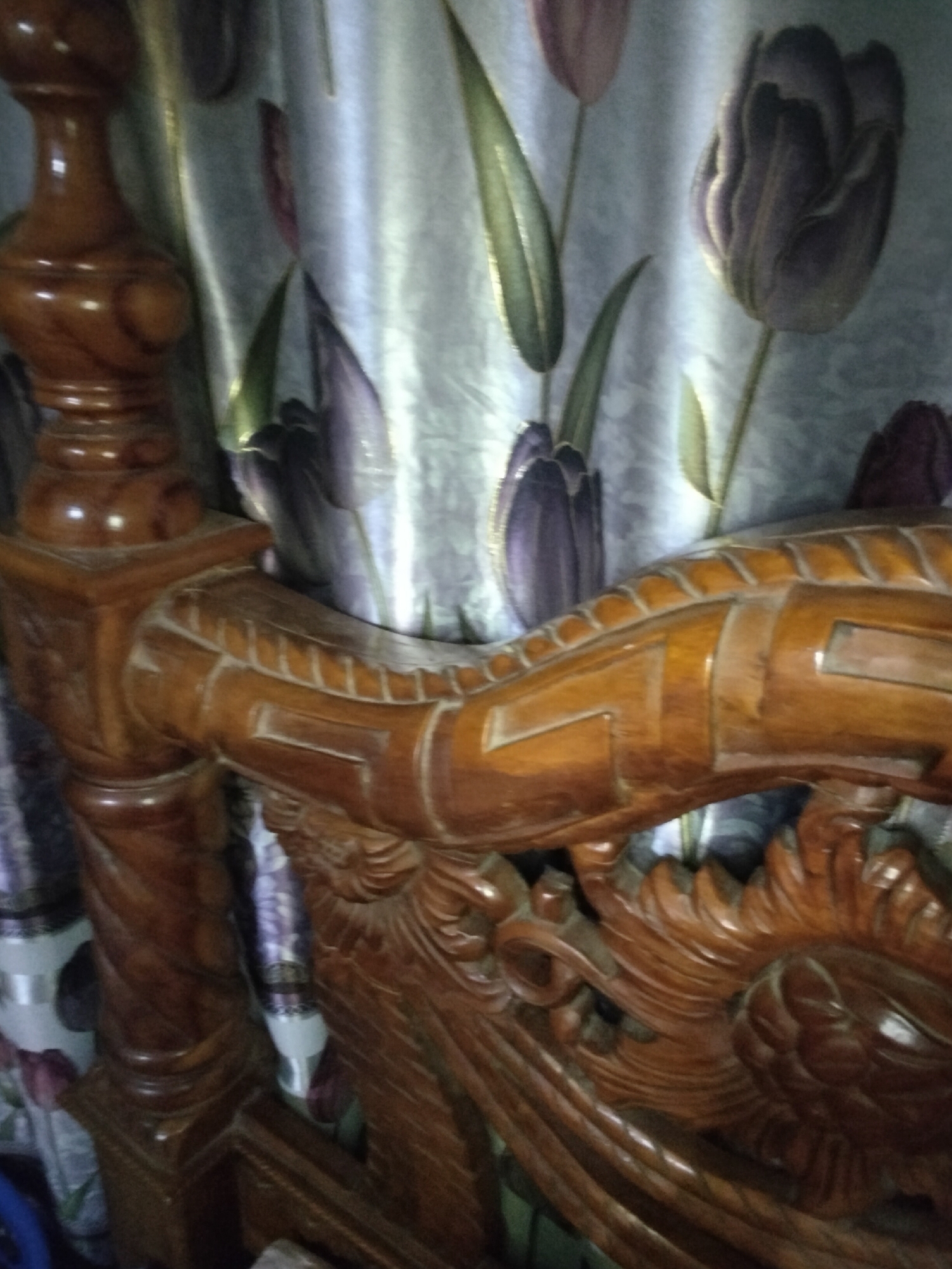 The image shows a close-up view of intricately carved wooden furniture, likely a headboard or footboard of a bed, in a room. The wood is a rich, polished brown and features elaborate scrollwork and possibly animalistic motifs, particularly on the right side of the frame where a carved face or mythical creature is visible. The backdrop consists of curtains with a printed pattern of stylized tulips in shades of purple and green against a light, possibly silver or grey, background. The lighting is dim, casting some shadows and highlighting the texture of the wood and the fabric. No people are visible in the image. The scene is static and appears to be an indoor setting, possibly a bedroom. The location within the city of Payagaboh, Indonesia, is not discernible from the visual information provided.