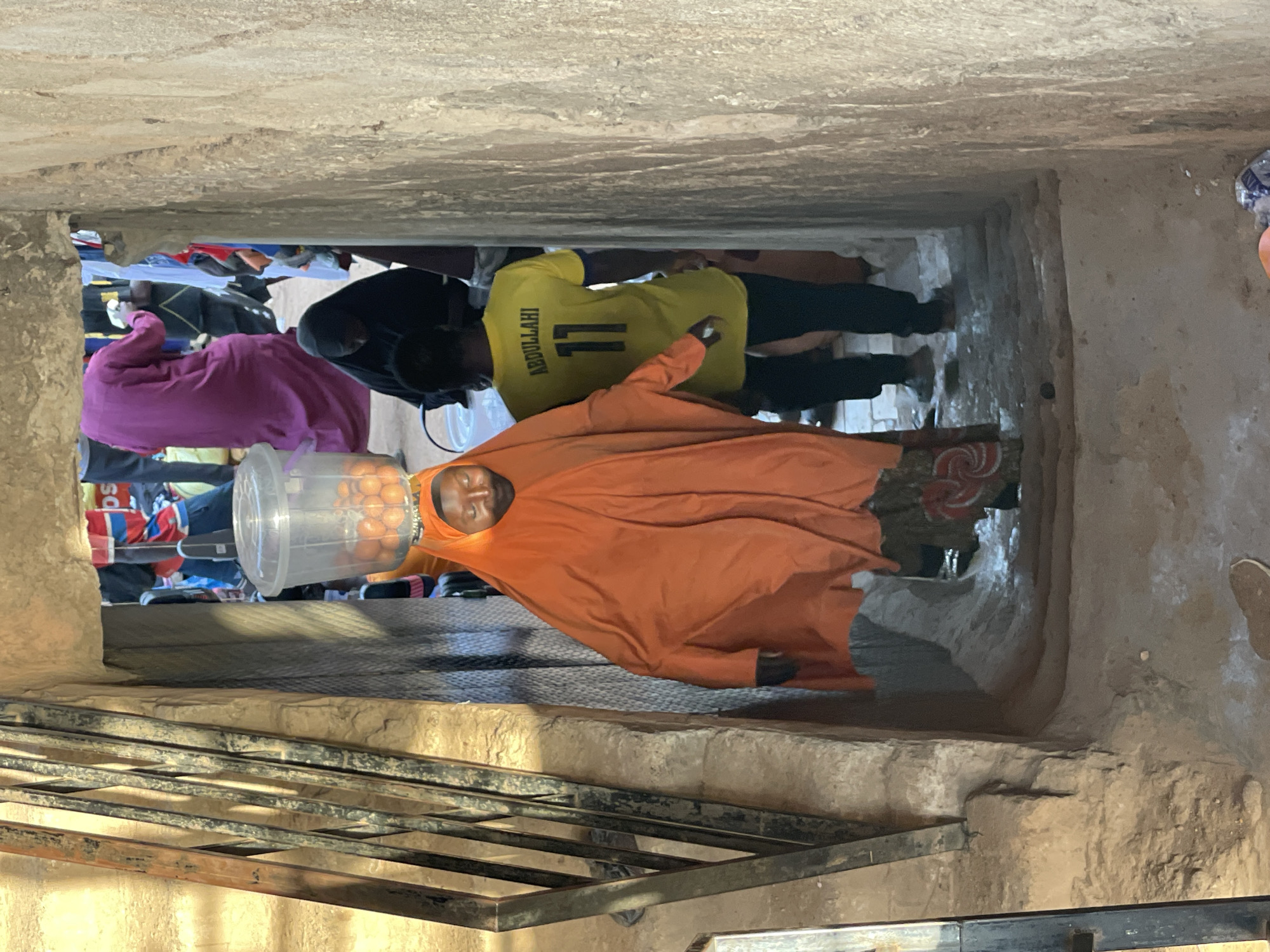 The image shows a woman wearing an orange hijab and carrying a plastic bucket full of oranges on her head. The woman is walking through a narrow, stone passageway in Bukuru, Nigeria. In the background, other people are visible, including a man wearing a yellow jersey with the name "ABDULLAHI" and the number "11" on the back. A metal grate and a stone wall are also visible.