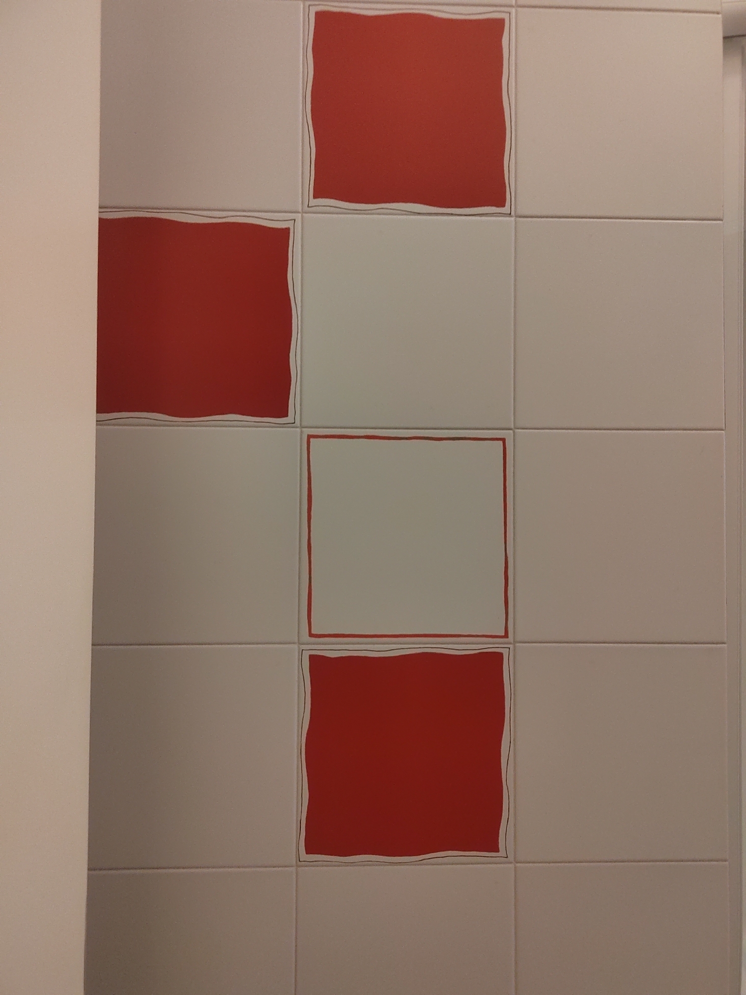 The image shows a wall covered in square tiles. There are alternating red and light gray tiles. The red tiles have a wavy, hand-drawn black outline. The wall is tiled in a grid pattern, with four rows and two columns visible. The lighting suggests an indoor environment. The image does not contain any people or specific landmarks that would indicate its location in Porto Recanati, Italy.