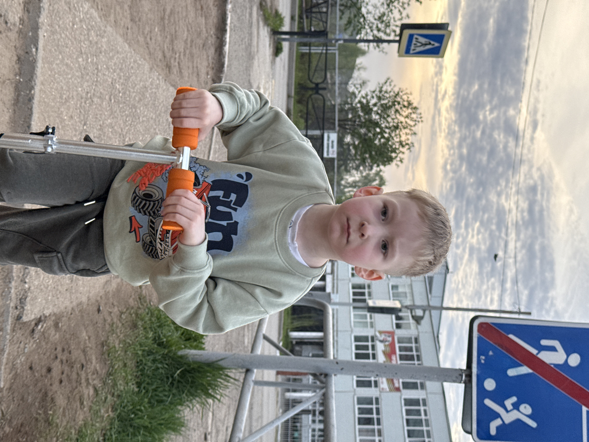 A young boy is standing on a scooter, holding the orange handlebars. He is wearing a light green sweater with graphics on it and grey pants. He is located near a sidewalk next to a patch of grass. In the background, there is a tall, multi-story building with many windows. There are two traffic signs visible: a pedestrian crossing sign and a sign indicating the end of a children's play area, located in the city of Chaikovsky, Russia. The sky is cloudy and bright.