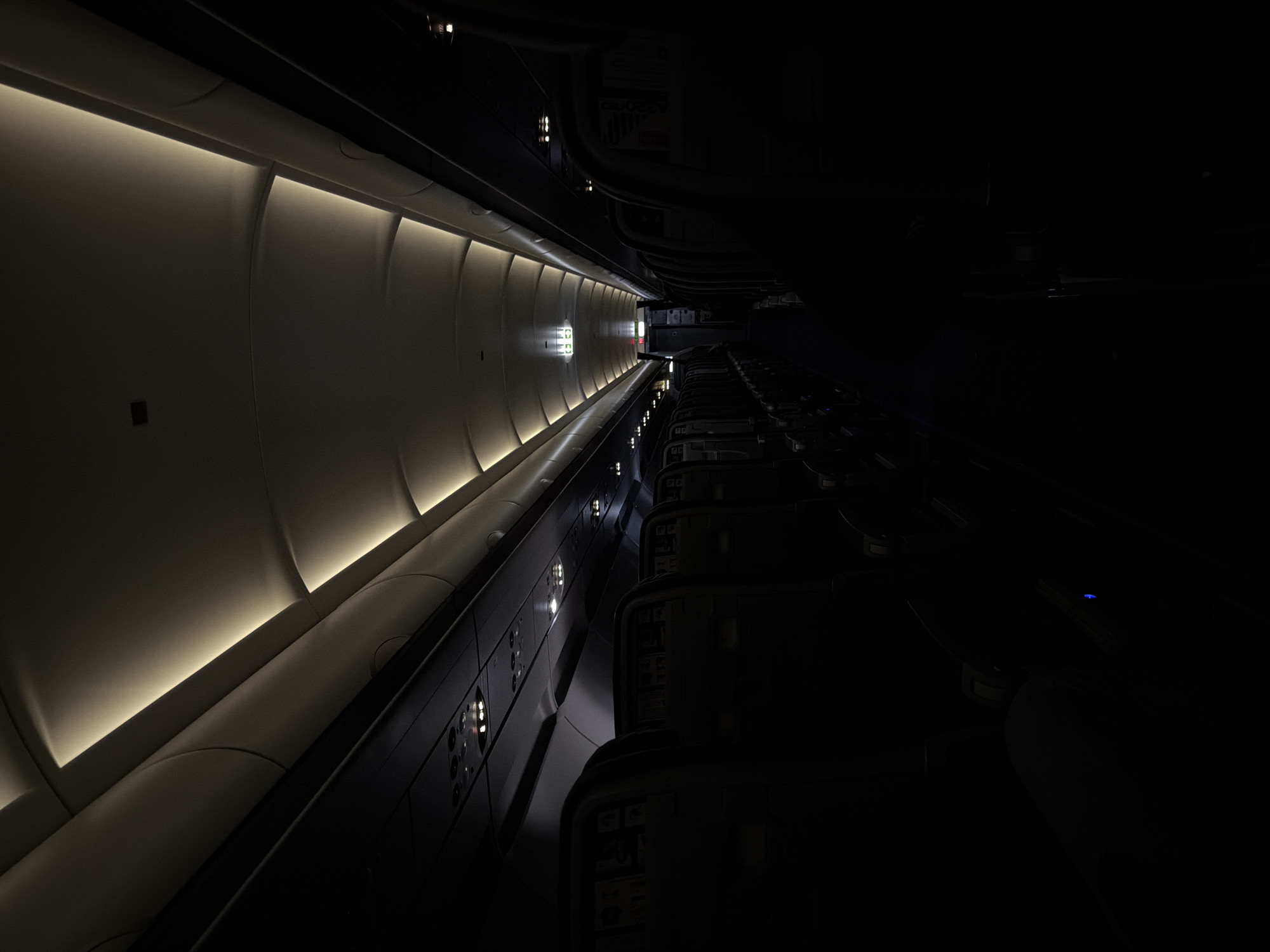 The image depicts the interior of what appears to be an aircraft cabin. On the left, the ceiling and upper sidewall are visible, illuminated by integrated light panels. The opposite side of the cabin shows rows of passenger seats in darkness. An emergency exit sign can be seen at the far end of the cabin. The specific location in Paiania Municipal Unit, Greece, is not determinable from the image alone.