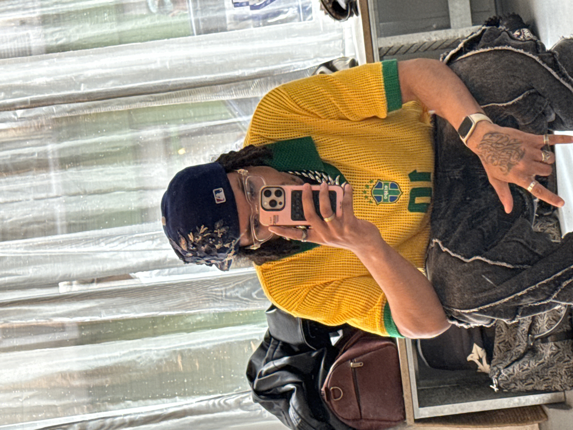 A person is taking a selfie in a mirror. The person is wearing a navy blue baseball cap with embroidery, a yellow Brazil soccer jersey with green trim and the number 10, eyeglasses, a silver chain necklace, multiple rings, and an Apple Watch. The person has dreadlocks and tattoos on their left hand. A black leather bag and a brown leather bag are visible near the bottom of the mirror. The background appears to be a window or a glass partition with a textured pattern. It is not possible to determine if the location is in Paris, France from the image alone.
