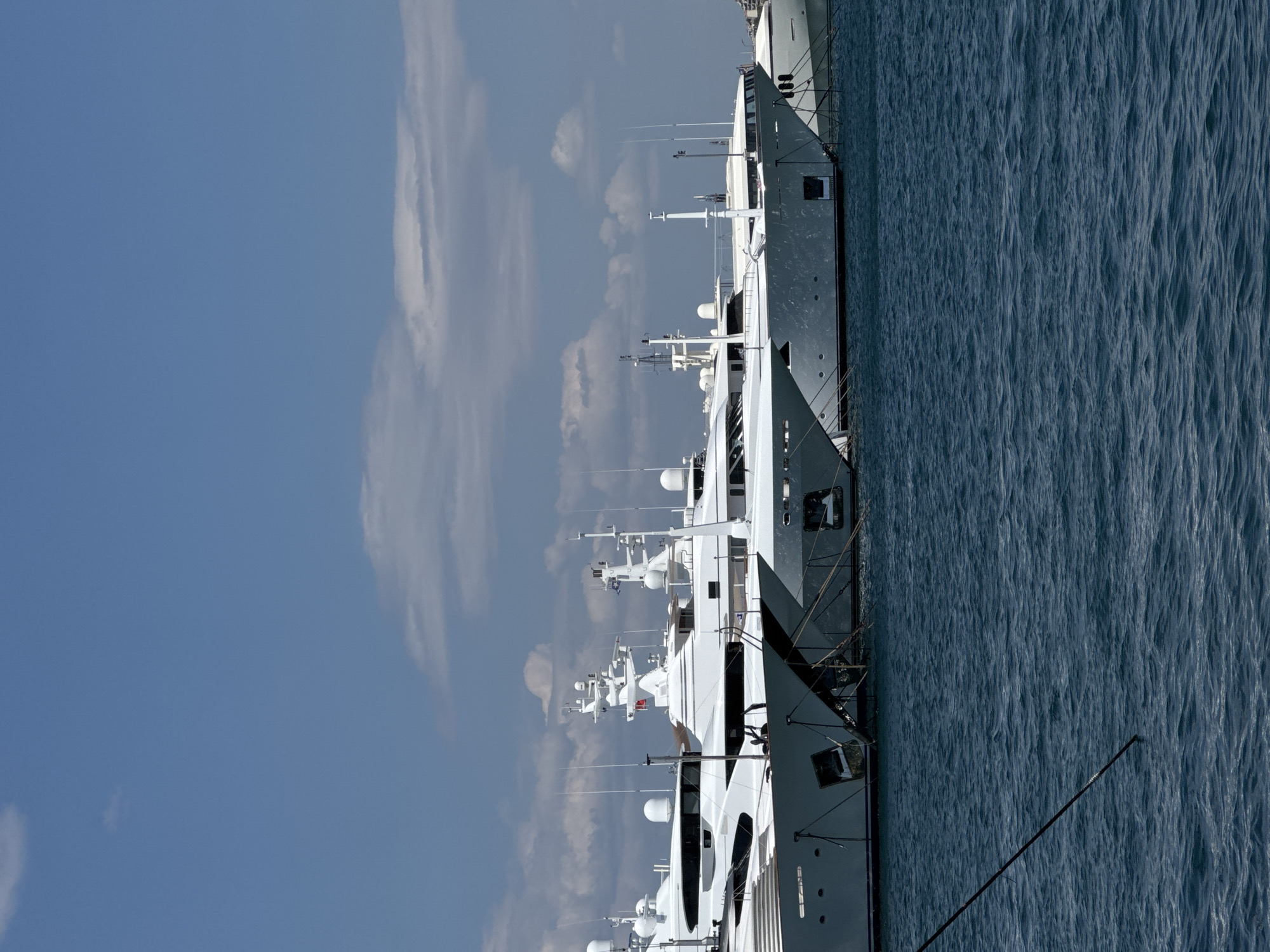 The image shows several large white yachts docked in a harbor under a partly cloudy blue sky. The yachts are lined up side-by-side, with their bows facing the viewer. They are all modern and luxurious, with sleek lines and multiple decks. The water is a deep blue and appears calm, with some gentle ripples. In the foreground, a dark rope extends diagonally across the frame, disappearing into the water. The background is mostly sky, with some scattered white clouds. The location is the port of Moschato, Greece.