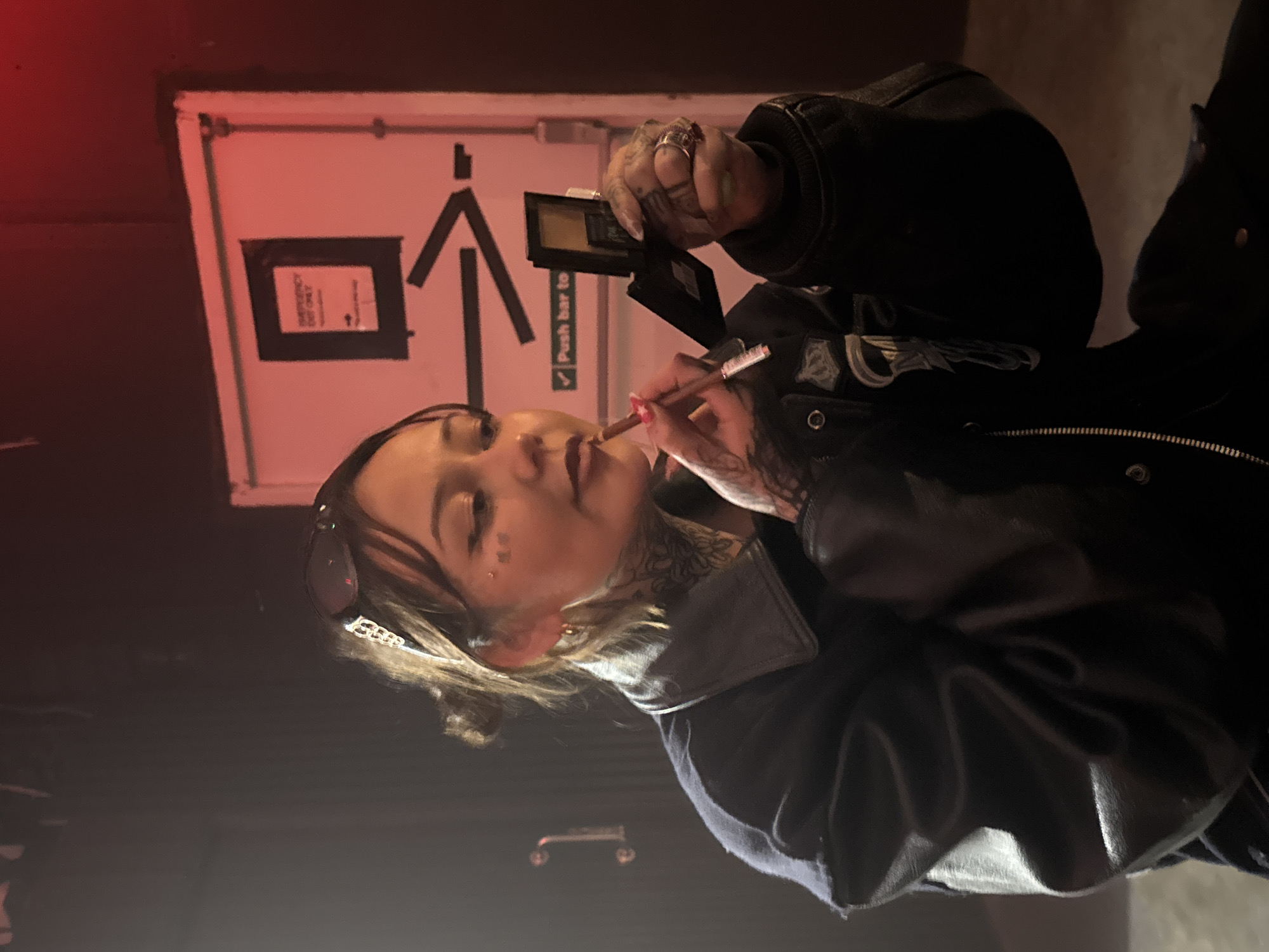 A woman with visible tattoos is applying makeup, holding a compact in her left hand and a brush in her right. She has sunglasses pushed up on her head and a decorative hair clip. The setting appears to be indoors, possibly backstage at a venue. A door is visible behind her with an arrow pointing upwards and the words "Push bar to" and a sign saying "Emergency Exit Only". The lighting is dim and has a red hue. This image was likely taken in London, United Kingdom, based on the style of signage and the urban aesthetic.