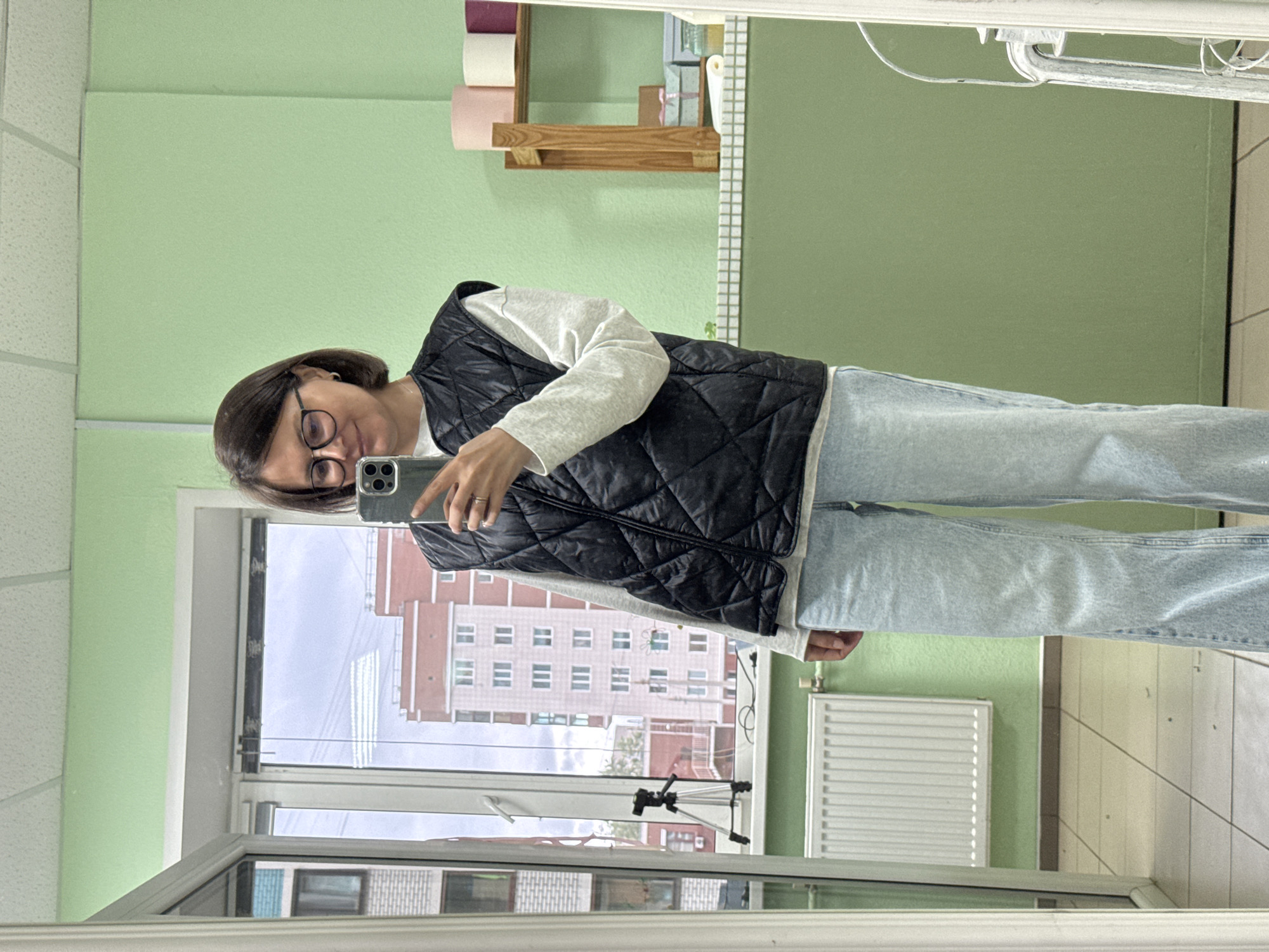 An adult female is depicted taking a mirror selfie in an indoor space located in the city of Izhevsk, Russia. The woman, with short dark brown hair and wearing round-framed glasses, smiles faintly while holding a grey smartphone with a multi-lens camera in her right hand to capture her reflection. She is dressed in a white long-sleeved top, a black quilted sleeveless vest, and light blue wide-leg jeans. A ring is visible on her right ring finger, and a small hoop earring is present in her left ear.
The interior features light green painted walls, a white acoustic tile drop ceiling, and a light-colored tiled floor. A large window with white frames occupies the left side of the scene, through which an overcast sky and several multi-story residential buildings, including one prominent light brown/orange structure, are visible. A black tripod stands on the windowsill, and a white radiator is located below the window frame on the far left. On the right side of the mirror's reflection, a wooden shelf unit displays rolls of pink, white, and purple material. Below the shelf, on a white-tiled ledge, small decorative boxes are arranged. A white-painted metal chair frame is partially reflected on the far right edge of the mirror.