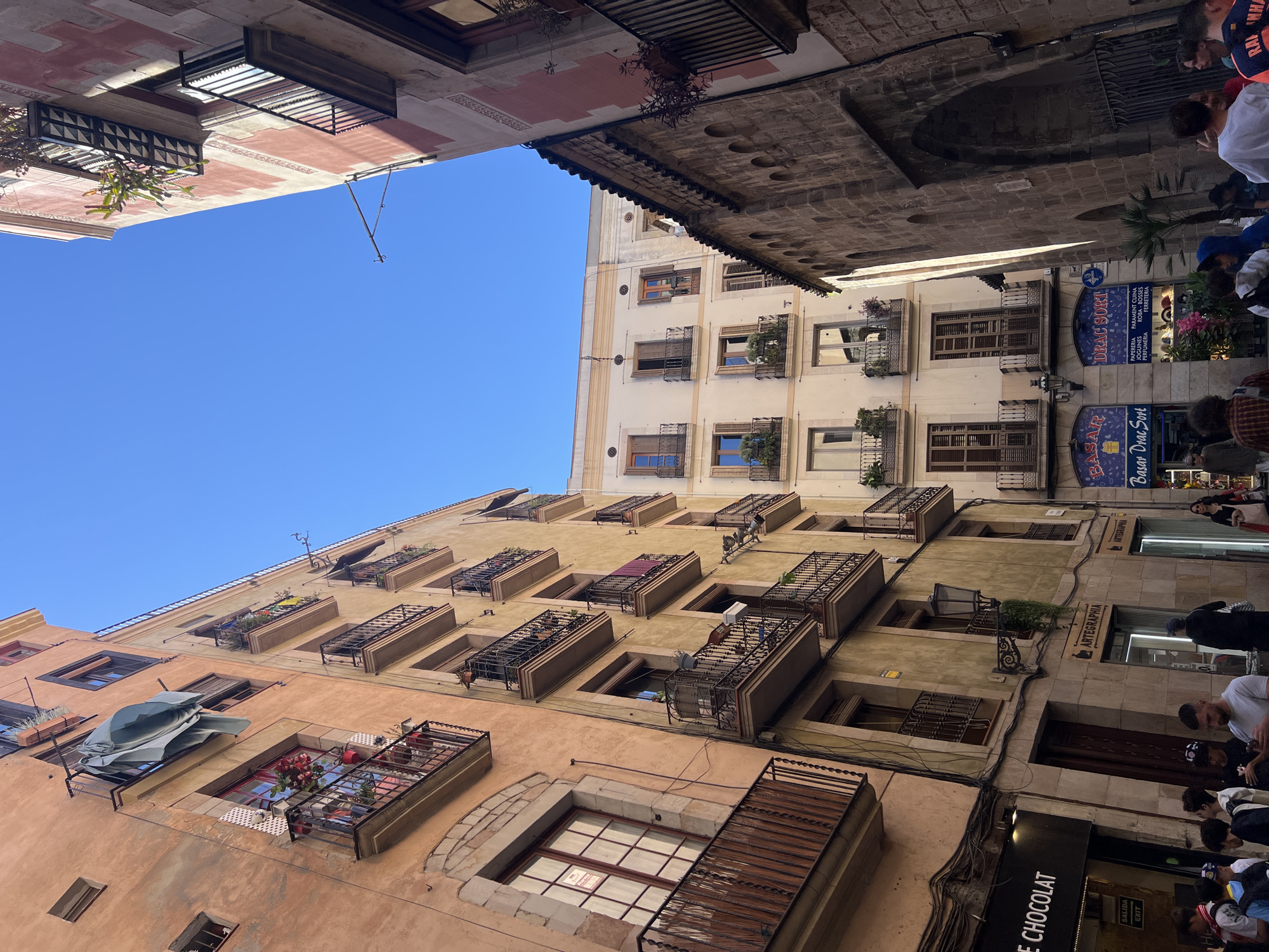 The image depicts a narrow street in Barcelona, Spain. The shot is angled upwards, capturing the facades of buildings lining the street and a patch of clear blue sky. The buildings are multi-storied with numerous windows and wrought-iron balconies, many adorned with flowers or plants. The buildings’ exteriors exhibit a range of colors, from pale yellow to orange and beige, suggesting an old and lived-in urban environment.
On the right side of the image, there are commercial establishments, identifiable by signs and storefronts. A store labeled "ARTEGRAPHIA" is visible, along with signs indicating shops like "DRAC SORT" and "Basar Drac Sort," which appear to sell stationery, toys, and perfume. People can be seen in the foreground, suggesting pedestrian traffic in the street. Cables and wires run along the buildings, adding to the urban texture. The overall impression is of a vibrant, historic street with a mix of residential and commercial activity.