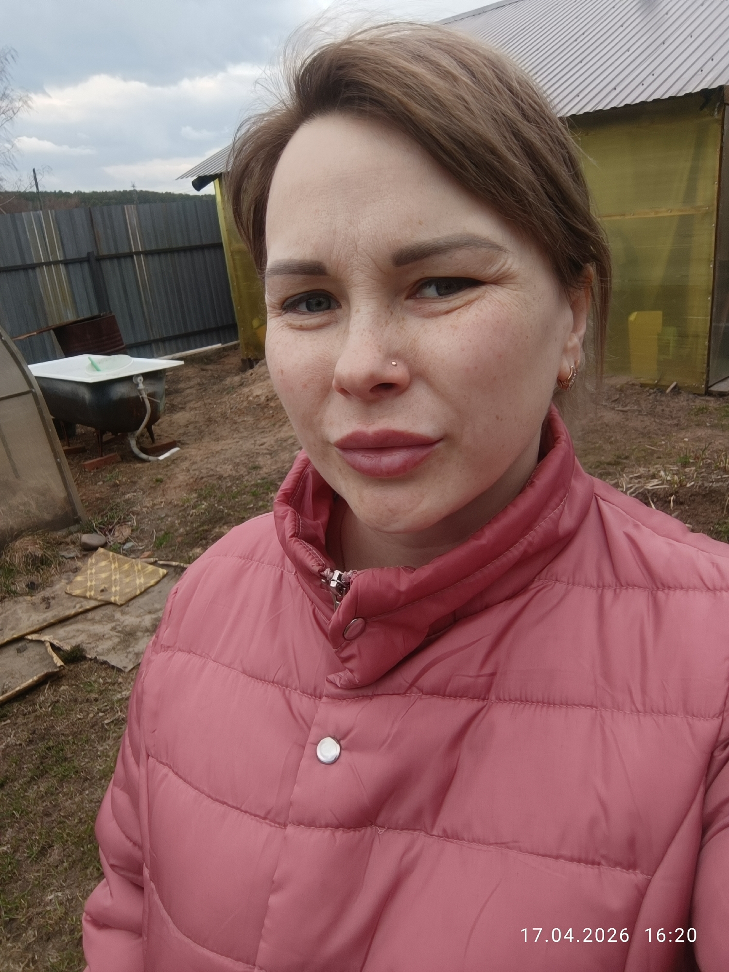 An outdoor selfie captures a woman in her 30s or 40s, looking directly at the camera with a slight, confident smirk. She has light brown hair, visible crow's feet around her eyes, and freckles across her nose and cheeks. Her lips are slightly parted and enhanced with light pink lipstick. A small silver stud adorns her right nostril, and she wears small hoop earrings. She is dressed in a rose-pink puffer jacket featuring a high collar and silver snap buttons.
The background reveals a rustic outdoor setting, likely a backyard or rural plot in Chaikovsky, Russia. The sky is overcast with light gray clouds. A tall, dark corrugated metal fence runs horizontally across the mid-ground, with trees and distant vegetation visible beyond it. To the right of the woman, a small structure with a corrugated metal roof and yellow, translucent walls—possibly a greenhouse or shed—is present. To her left, near another smaller greenhouse-like structure, an old, dark-colored bathtub on a stand holds a white basin and has a hose attached. A rusty barrel is also in the vicinity. The ground is covered with dry grass, patches of dirt, and some scattered debris, including a yellow patterned mat.
The bottom right corner of the image displays a time stamp: "17.04.2026 16:20", indicating the date and late afternoon time.