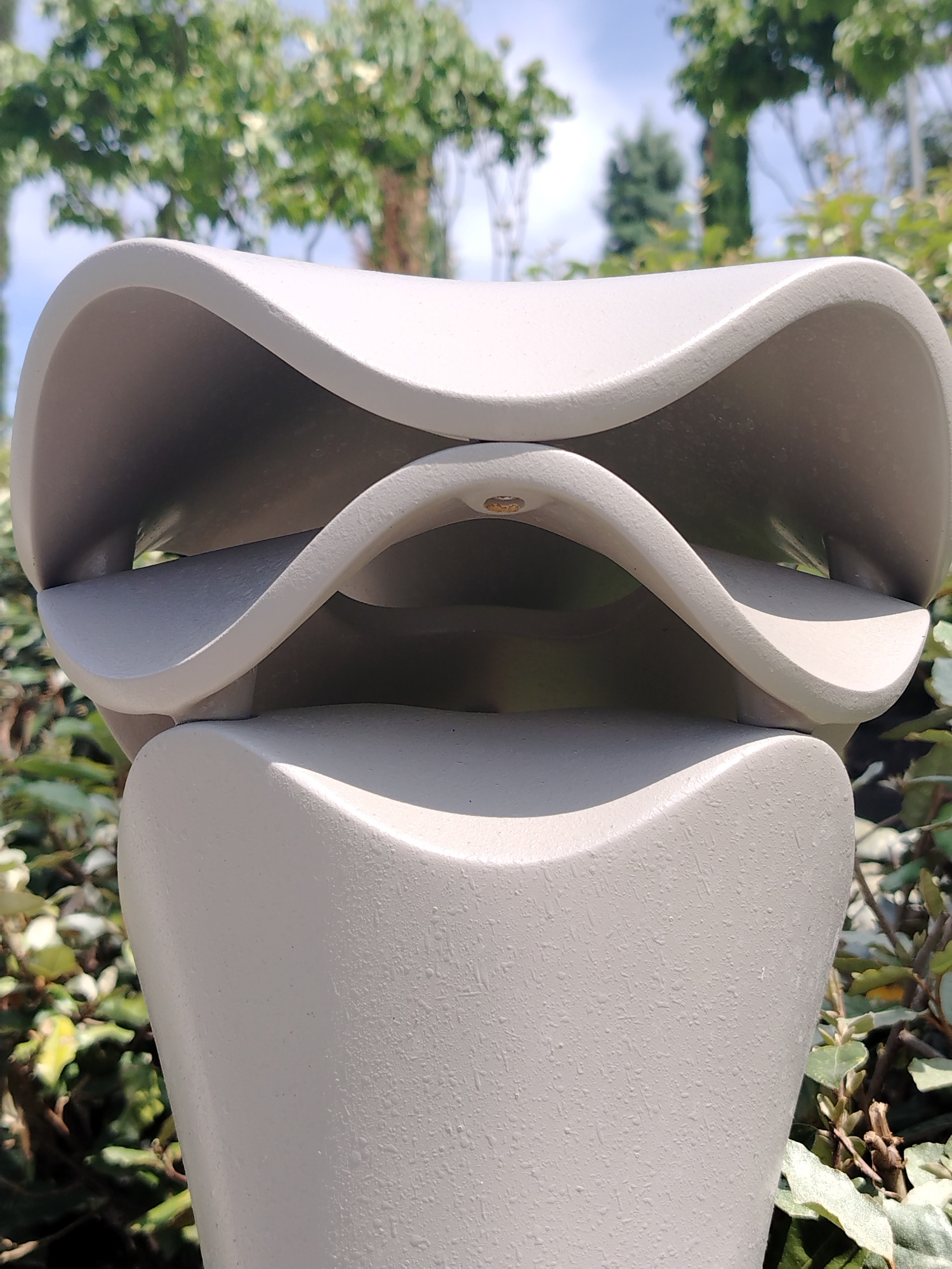 The image shows a light gray, wavy decorative sculpture placed outdoors in front of a backdrop of green foliage and trees under a bright sky. The sculpture appears to be made of a textured material and has several curved layers. The lighting suggests it is daytime. Without further information or visual cues, the specific location within the city of Krasnodar, Russia cannot be determined.