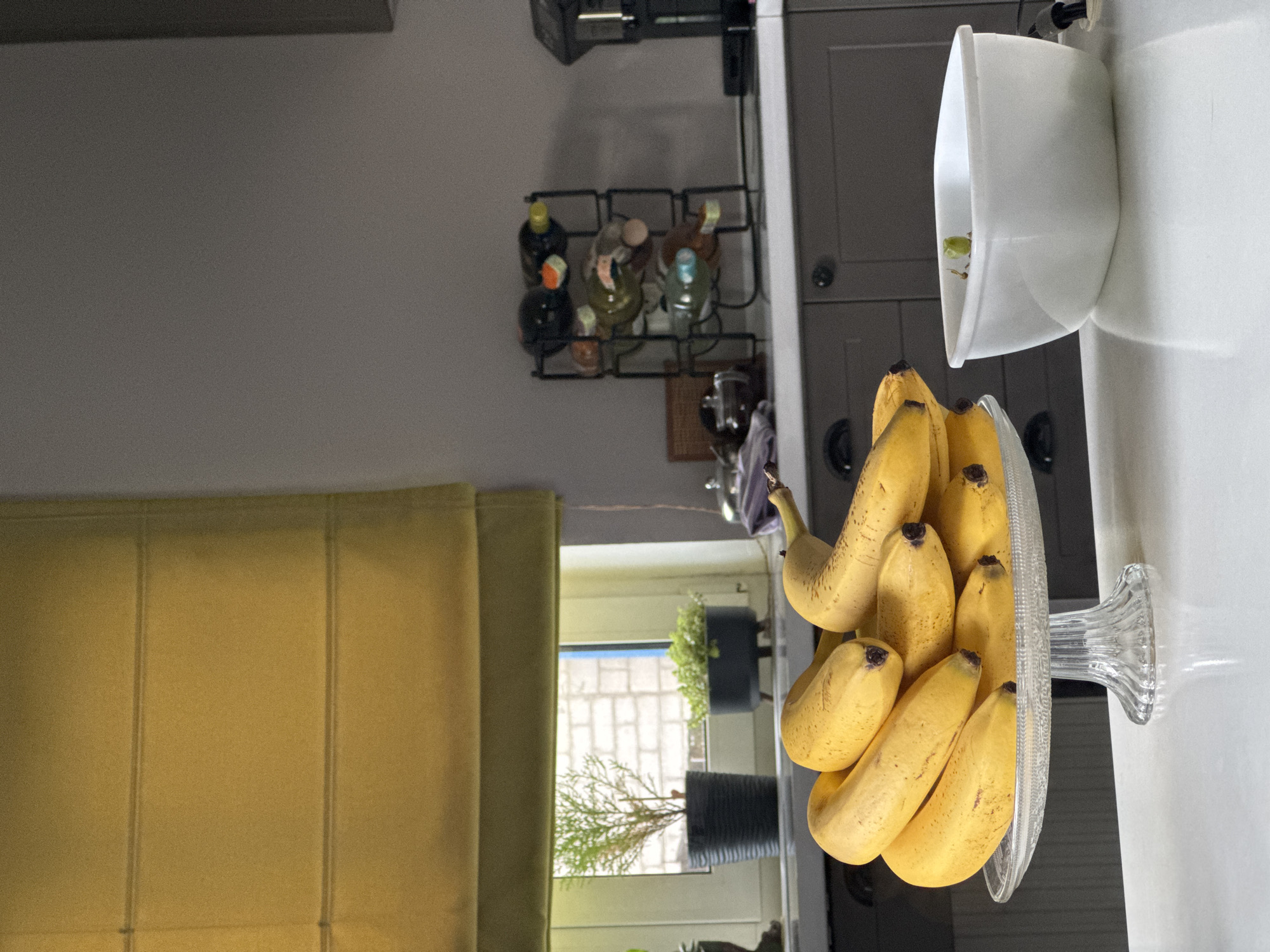 The image depicts a kitchen scene with a bunch of ripe bananas in a clear glass fruit bowl on a white countertop. Behind the bananas, a window with yellow roman blinds is visible. Two potted plants sit on the windowsill. Further back on the countertop, a black metal wine rack holds several bottles of wine. A white square container is positioned to the right of the bananas. The kitchen cabinets are dark grey. The scene appears to be set in a domestic environment. There is no information in the image to suggest it is located in Kremenchuk, Ukraine.