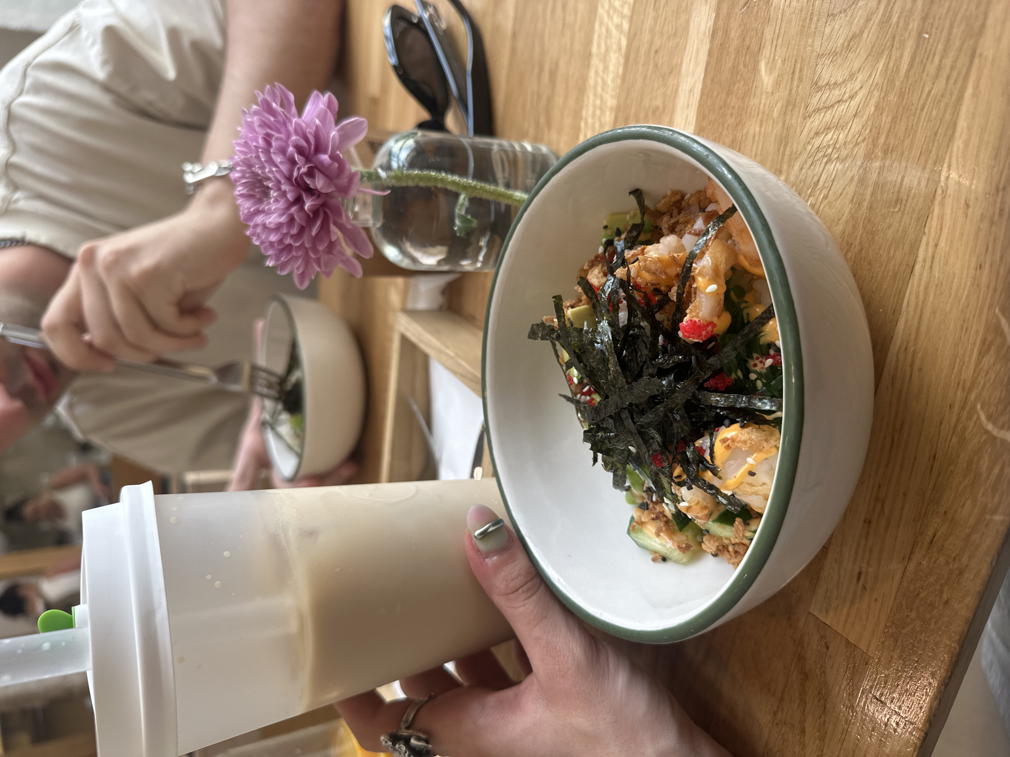 A close-up, high-angle shot shows a hand holding a large, clear plastic cup with a white lid and a straw. The cup contains a light-colored beverage with ice. In the foreground, a white bowl with a green rim sits on a wooden table. The bowl is filled with a poke bowl containing rice, shrimp, avocado, cucumber, and seaweed. A small glass vase with a purple flower is placed behind the bowl, and a pair of sunglasses rests on the table further back. In the background, two people are visible, one of whom is eating from a bowl with a fork. The setting appears to be a cafe or restaurant in Moscow, Russia, indicated by the visible signage outside the establishment.