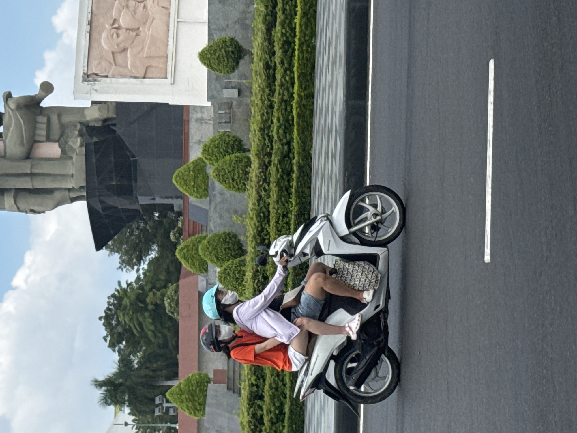 Two young Vietnamese women are riding on a white scooter on a paved road in Phan Thiết, Vietnam. The woman in the front is driving the scooter and wearing a light purple long-sleeved shirt, blue helmet, and a face mask. She has dark hair and is wearing denim shorts. The woman behind her is wearing an orange t-shirt, a red and black helmet, and a face mask. She has dark hair and is wearing white shorts. Both women are wearing masks. The scooter is traveling on the right side of the road, which has a white line marking the lane. In the background, there is a large, modern monument with sculpted figures and a white relief panel. There are also manicured bushes and trees, and a cloudy sky. The image is taken in daylight.