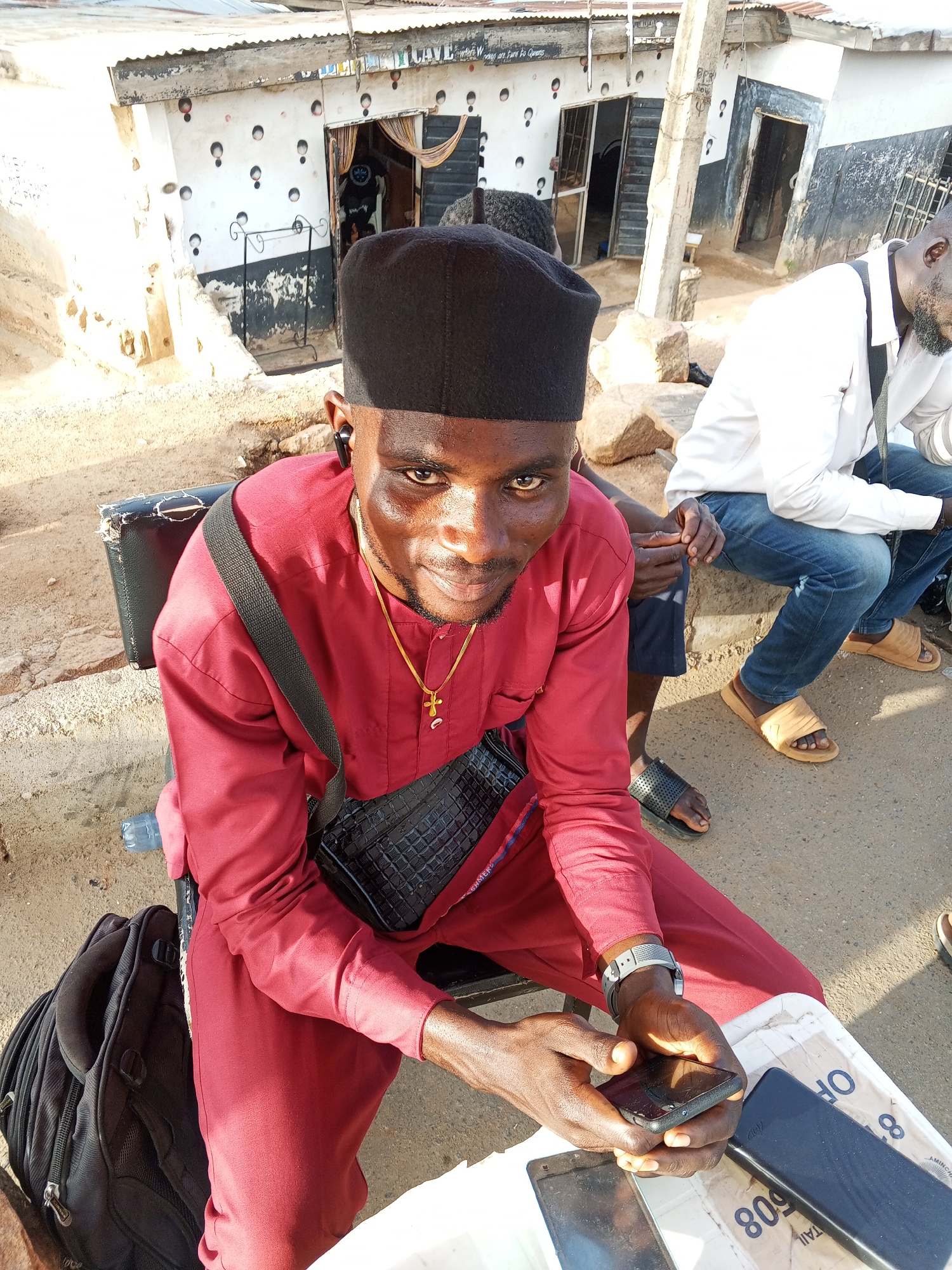 A man in a red shirt and black cap sits on a chair, looking at the camera while holding a smartphone. He wears a crossbody bag and a gold necklace with a cross. Three phones rest on a white surface in front of him. Another man sits on a concrete structure in the background, wearing a white shirt, blue jeans, and sandals. The setting appears to be outdoors in Gwong, Nigeria, near a building with a metal roof and decorative painting on the wall.
