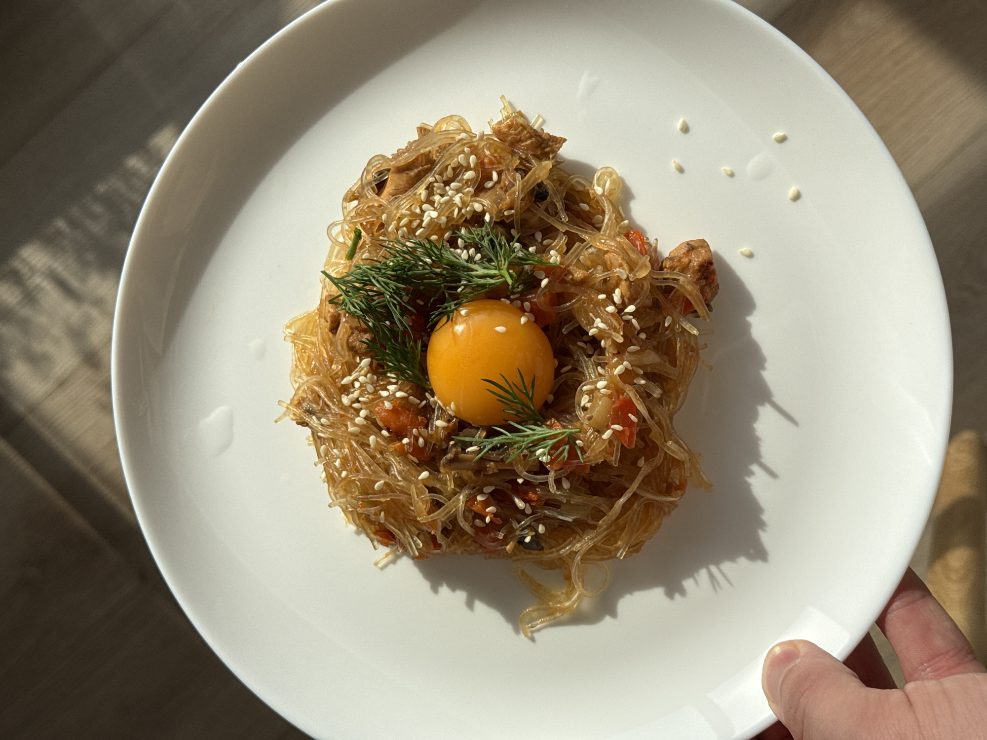 The image shows a white plate with a mound of cellopane noodles topped with sesame seeds, dill, and a bright yellow egg yolk. The plate is being held by a hand in the bottom right corner. The location is unspecified.