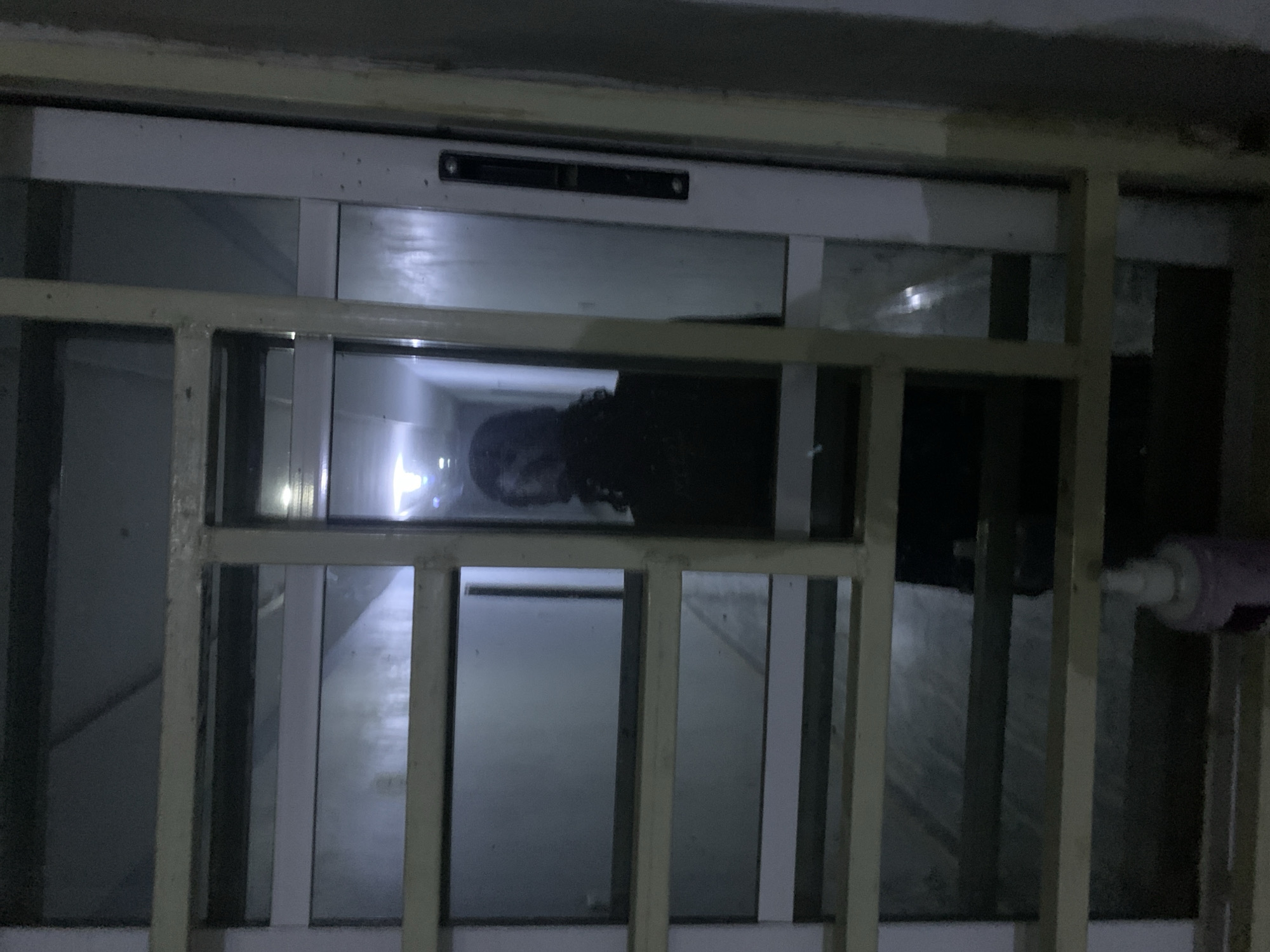 The image captures an interior view looking through a light-colored, grid-patterned security barrier and a glass door or partition within a structure in the city of New Stadium, Nigeria. In the immediate foreground, the metallic or painted security grid occupies the central and left portions of the frame. Below the grid, on a surface, a small, light-colored bottle with a white pointed dispenser cap is visible.
Behind the security grid is a glass panel framed by white vertical and horizontal elements. The glass reflects a dark-clad individual with dark, potentially curly hair, visible from the chest up on the right side of the frame. This appears to be a reflection of the photographer or a person standing directly in front of the glass.
Beyond the glass, a dimly lit, narrow corridor or hallway is visible, extending into the background. The walls of the corridor appear light-colored. A prominent bright light source is present at the far end of this corridor, illuminating the ceiling and creating a distant focal point. The floor of the corridor appears to be a uniform, dark surface. The overall lighting is low, with the distant corridor light being the primary illumination beyond the glass. The surfaces of the grid and door frames show minor wear and specks.