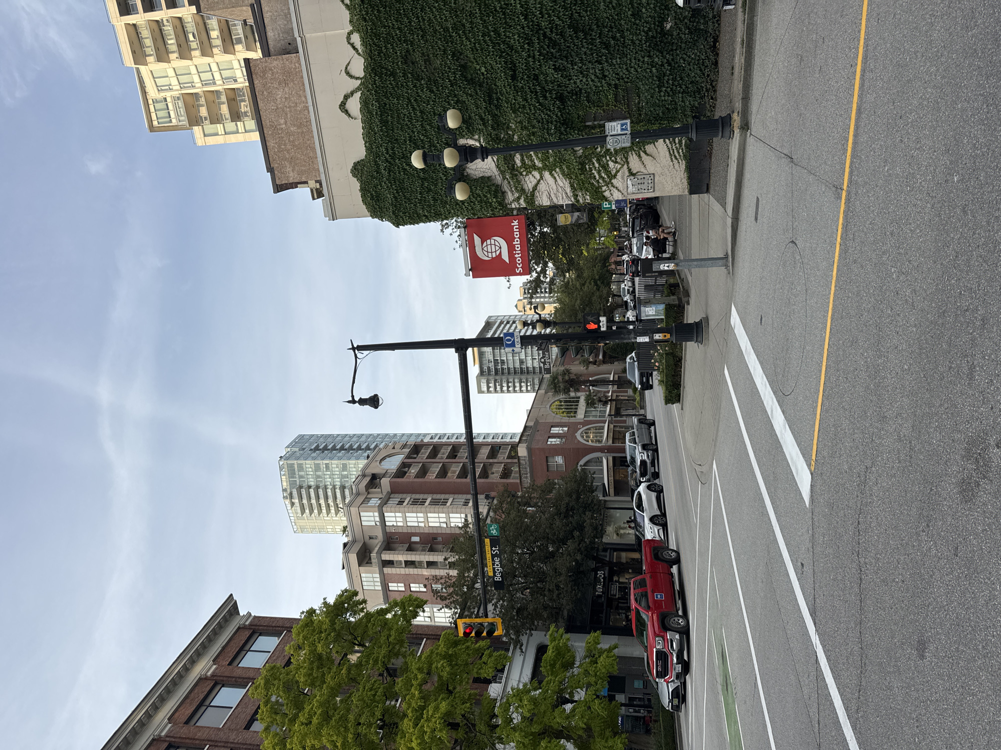 The image captures a street scene in New Westminster, Canada, on a cloudy day. The viewpoint is from the middle of a street, looking towards a bustling urban intersection. The foreground features a paved road with clear lane markings, including white crosswalk stripes and a yellow line. A red GMC pickup truck is prominent on the left side of the road, stopped at a traffic light.
The intersection is marked by a traffic light displaying a yellow signal. A street sign indicating "Begbie St." is visible, along with a bicycle lane symbol. Multiple buildings line the street, displaying a mix of architectural styles. On the left, a red brick building with white trim stands tall, partially obscured by lush green trees. Across the street, a modern, light-colored building covered in dense green ivy dominates the right side of the frame. A red "Scotiabank" sign is mounted on this ivy-covered building.
Further down the street, several other buildings, including high-rise residential towers, are visible against a pale blue sky with scattered clouds. Various vehicles, including cars and SUVs, are either parked or in motion on the street. Pedestrians can be seen on the sidewalks. The overall environment suggests a lively city atmosphere with a blend of older and contemporary architecture.