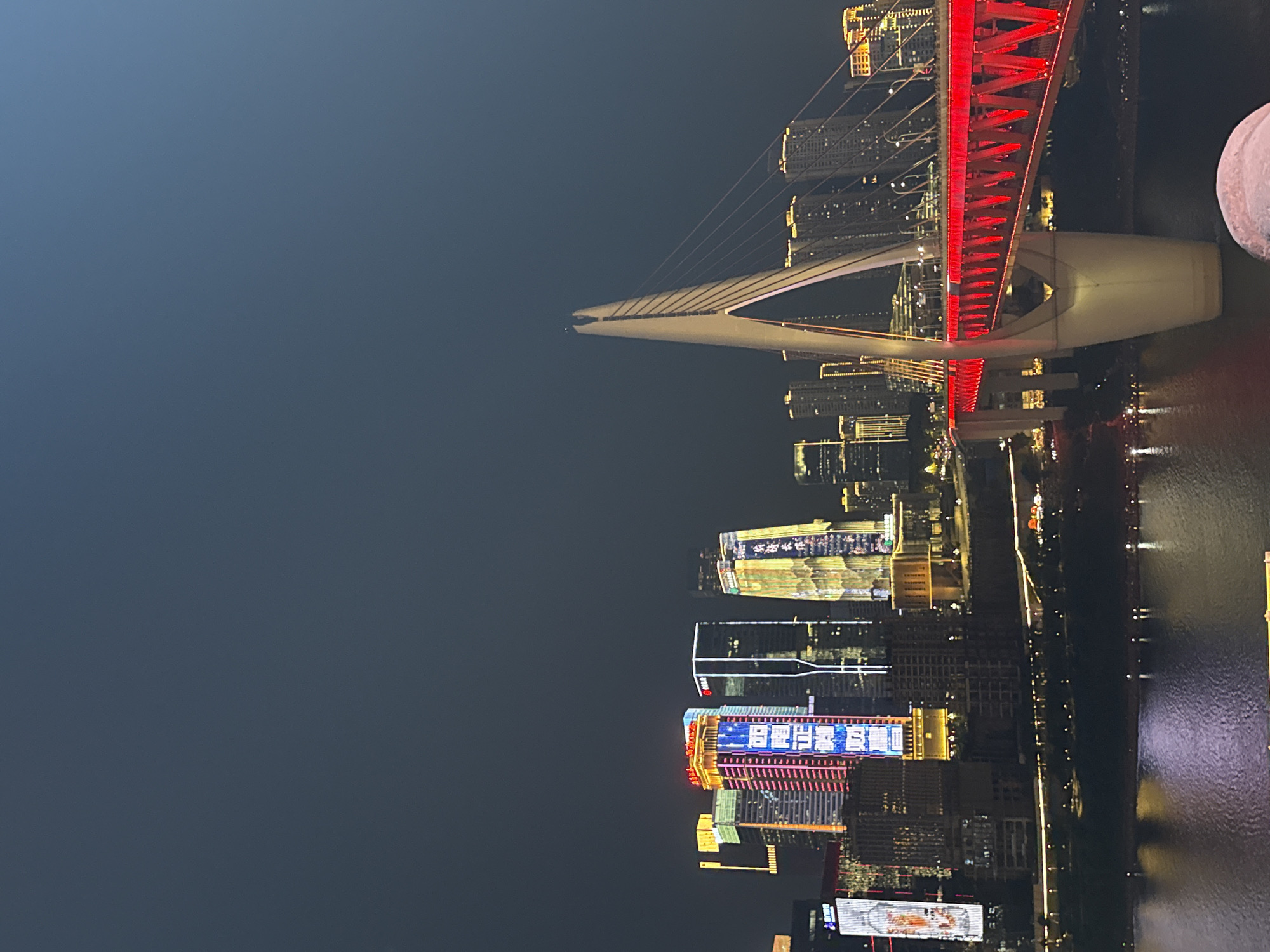 The image captures a night-time cityscape in Yuzhong District, China, dominated by the illuminated Hongyadong Bridge and a skyline of skyscrapers. The Hongyadong Bridge, a prominent cable-stayed bridge, is brightly lit with vibrant red lights along its deck and illuminated white towers, with cables extending upwards. In the background, a cluster of modern skyscrapers stands tall, adorned with colorful, dynamic LED displays and architectural lighting, with some buildings featuring vertical Chinese text and patterns. A wide river, reflecting the city lights, flows in the foreground. The sky is a deep, dark blue, characteristic of nighttime. No people are visible in the frame.