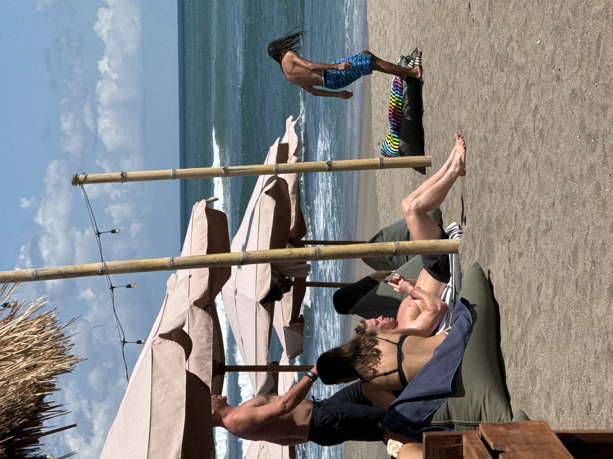 The image depicts a beach scene in North Kuta, Indonesia. In the background, a man with dreadlocks is performing a mid-air trick. In the foreground, three people are lounging under thatched umbrellas. One of them is lying on a cushioned seat with a towel.
