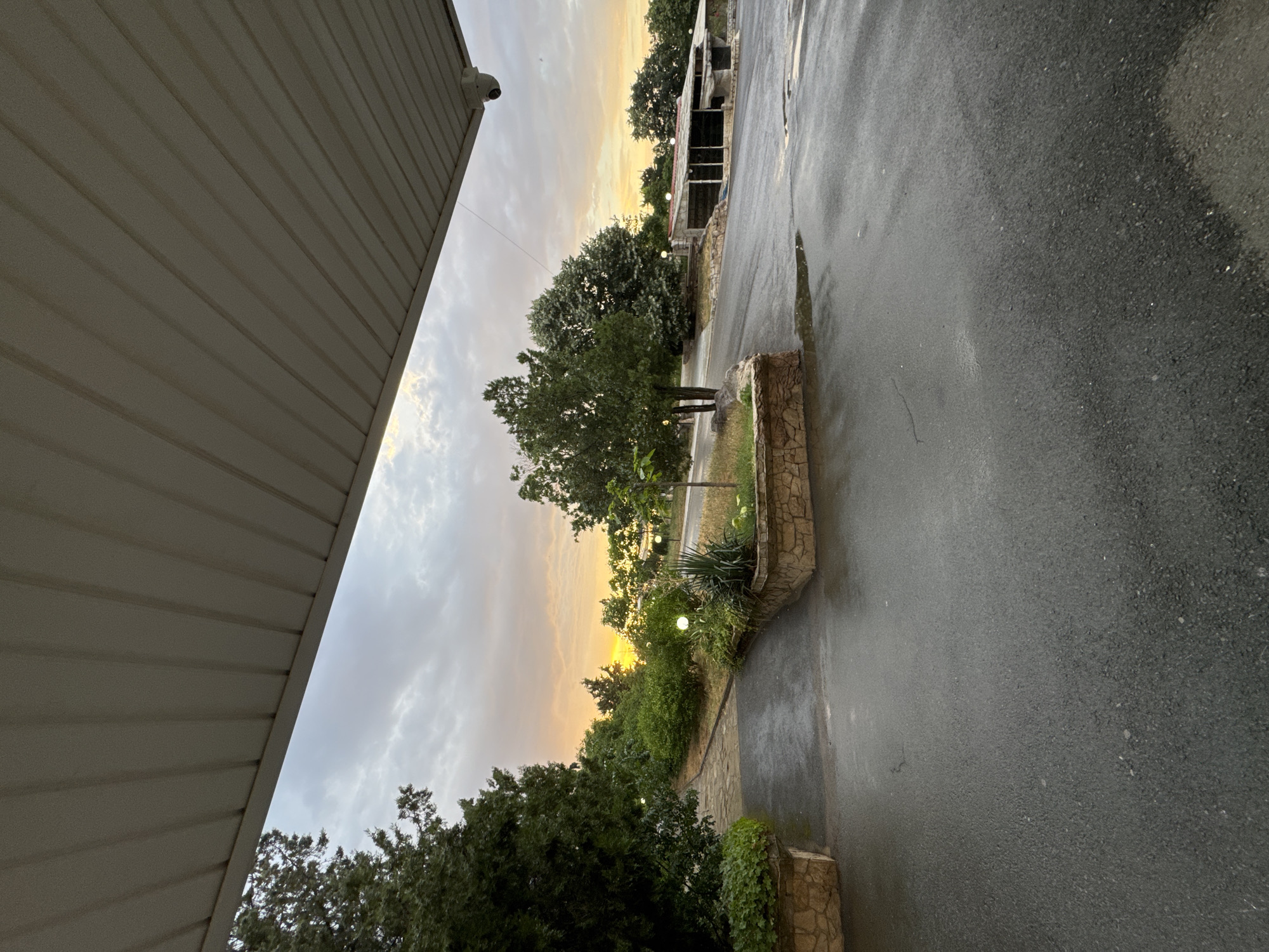 The image depicts an outdoor scene at dusk, likely in Sredets, Bulgaria. The viewpoint is from under a covered structure, possibly a porch or overhang, with a textured, beige ceiling visible in the foreground. To the left, a white security camera is mounted. The scene transitions to a view of a wet asphalt road, indicating recent rain or damp conditions. The road is bordered by a low, stone-lined planter box filled with greenery and small plants. Beyond this, a path made of light-colored pavers winds through a garden area. Several trees with lush green foliage are scattered throughout the landscape, and a few round, illuminated garden lights are visible amongst the plants. In the background, a partially constructed or dilapidated building with an exposed framework stands amongst the trees. The sky is filled with dramatic clouds, and a warm, golden sunset is breaking through, casting a soft glow on the horizon. The overall mood is serene and atmospheric, capturing the transition from day to night. There is no visible text in the image.