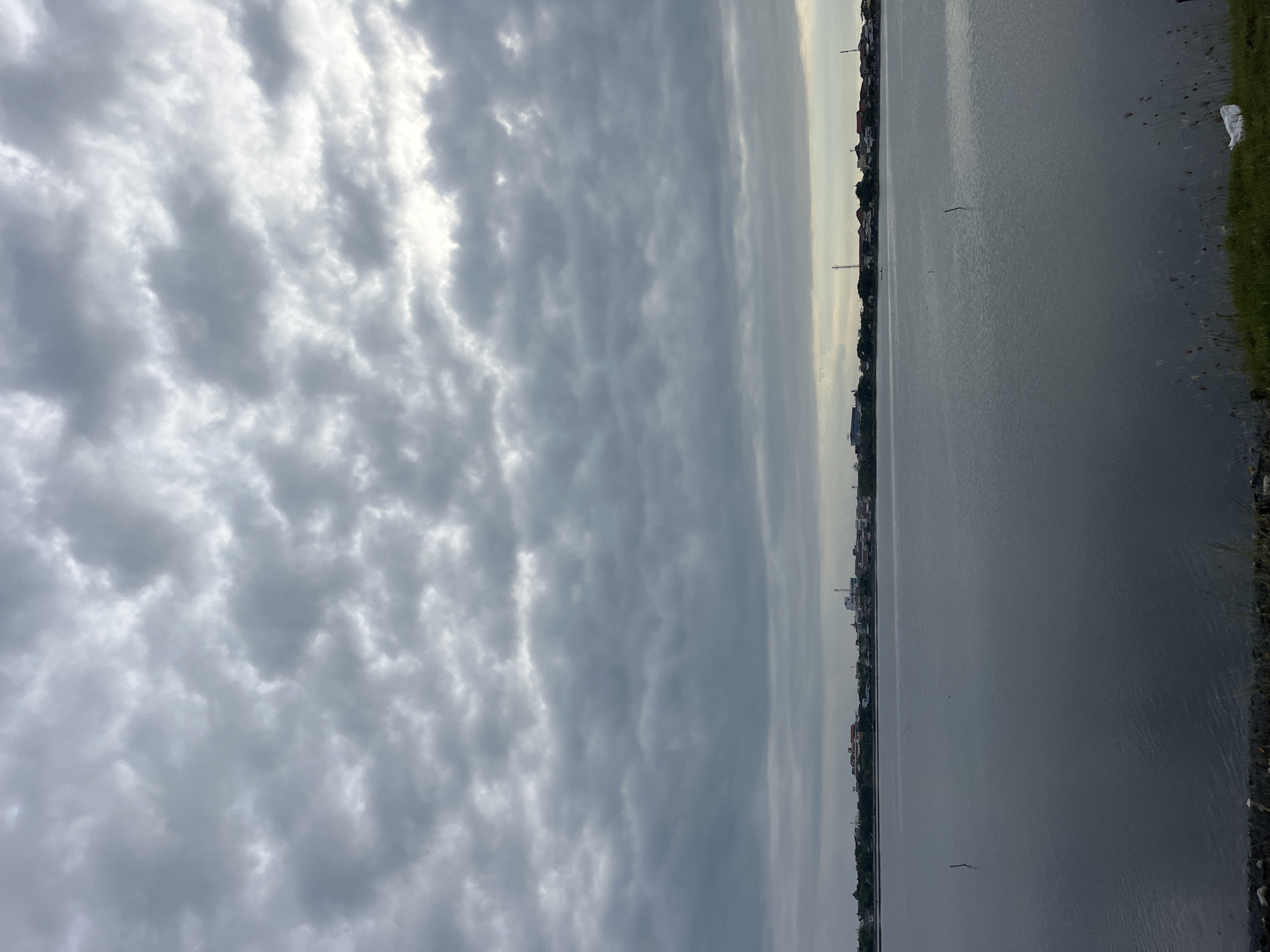 The image shows a waterfront scene in Lhokseumawe, Indonesia. The sky is filled with overcast clouds. A body of water stretches along the right side of the frame, its surface reflecting the sky. A cityscape is visible along the far side of the water, with buildings and vegetation lining the shore. The immediate shoreline is visible in the bottom right corner, with some grass and debris.
