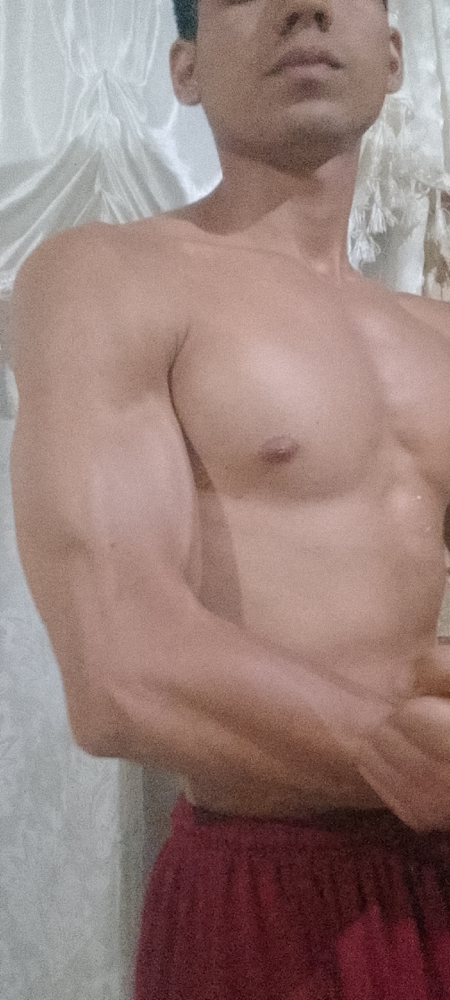 A shirtless man is posing indoors against a backdrop of white curtains. He is wearing burgundy shorts and has muscular arms and chest. The location is indeterminate, and there is no evidence to suggest the image was taken in Juan Griego, Venezuela.