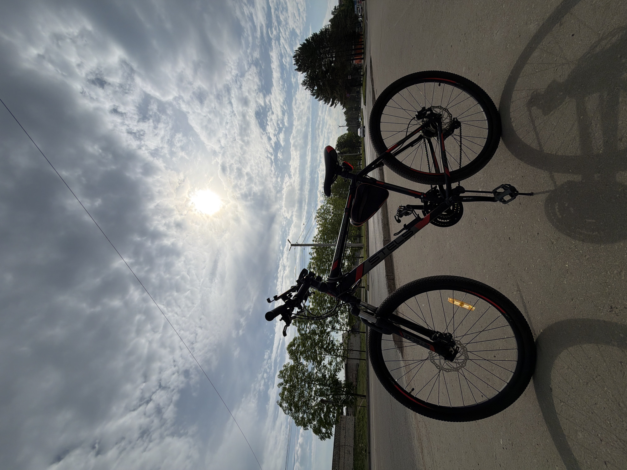 A black and red mountain bike lies on its side on a gray concrete surface, casting a shadow. The background is a sky with numerous white and gray clouds, and the sun is visible through the clouds. In the distance, trees and buildings are visible. The location is Birobidzhan, Russia.