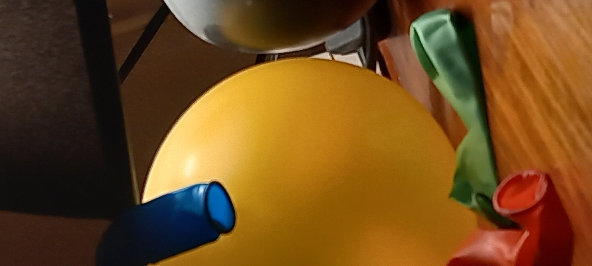 This close-up image captures an abstract arrangement of colorful objects, likely indoors in Kryvyi Rih, Ukraine. The most prominent feature is a large, vibrant yellow, rounded object, which appears to be an inflated balloon or a large ball, dominating the central and lower-right portion of the frame.
Extending from the lower-left edge and partially overlapping the yellow object is a blue cylindrical form, resembling an air pump nozzle or the neck of another balloon. At the top of the image, a segment of a white or light gray spherical object is visible, possibly another balloon or a reflective surface.
On the right side, two deflated balloons are present. A green, elongated, and crumpled balloon rests against a reddish-brown background, with a red, similarly deflated and crumpled balloon positioned below it. The background behind these elements features a textured, light brown or wooden surface with discernible vertical grain, while the left side of the image shows a darker, less defined surface, possibly fabric or a wall. Dark linear elements within the darker background might be parts of furniture or structural supports.
No people are visible in the image, nor is any text present. The collection of inflated and deflated balloons, along with the nozzle-like object, suggests a context of celebration or play, possibly capturing either the process of decorating for an event or its conclusion. The indoor setting provides no specific indicators of the time of day or weather conditions.