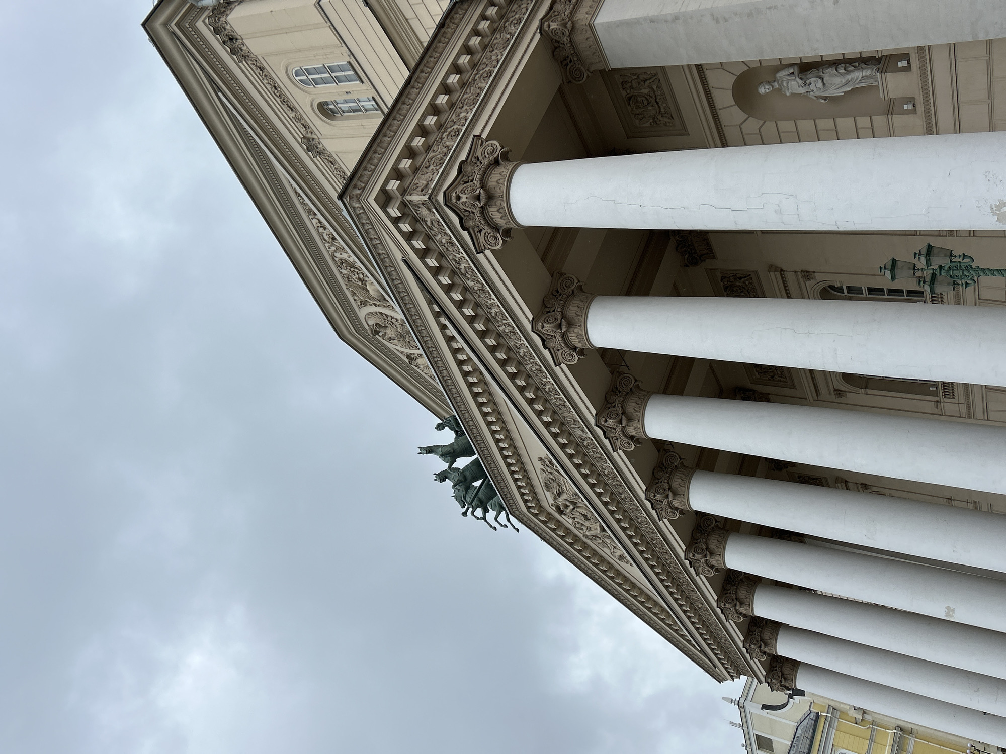 The image depicts a low-angle view of a classical building facade against a cloudy sky. The architectural details are prominent, featuring a row of white columns with ornate capitals. Above the columns, the building's entablature is adorned with intricate carvings and moldings. A statue of a chariot pulled by horses is visible on the roofline. In the background, portions of other buildings can be seen, partially obscured by the low angle. The overall tone of the image is subdued, reflecting the overcast weather conditions.