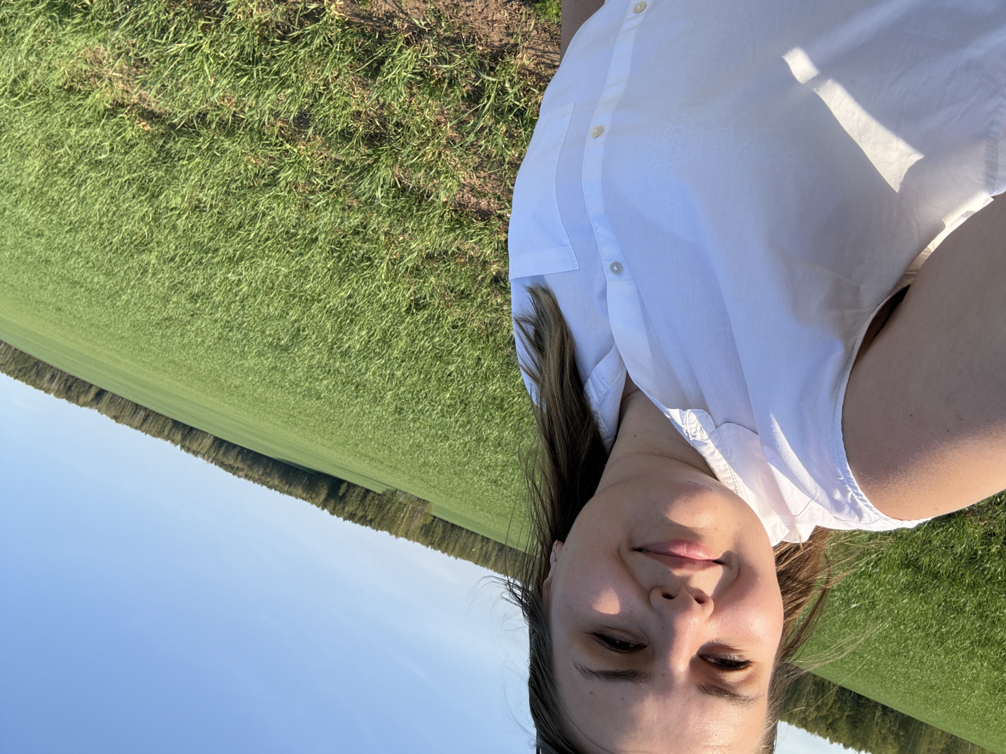 A person, likely female, is seen in a selfie. She has light skin, brown hair, and is wearing a white button-down shirt. The background features a green field and a strip of trees bordering the field. The sky is blue. The location appears to be an outdoor rural setting near the city of Шумиха, Russia. The photo appears to be taken during the day.