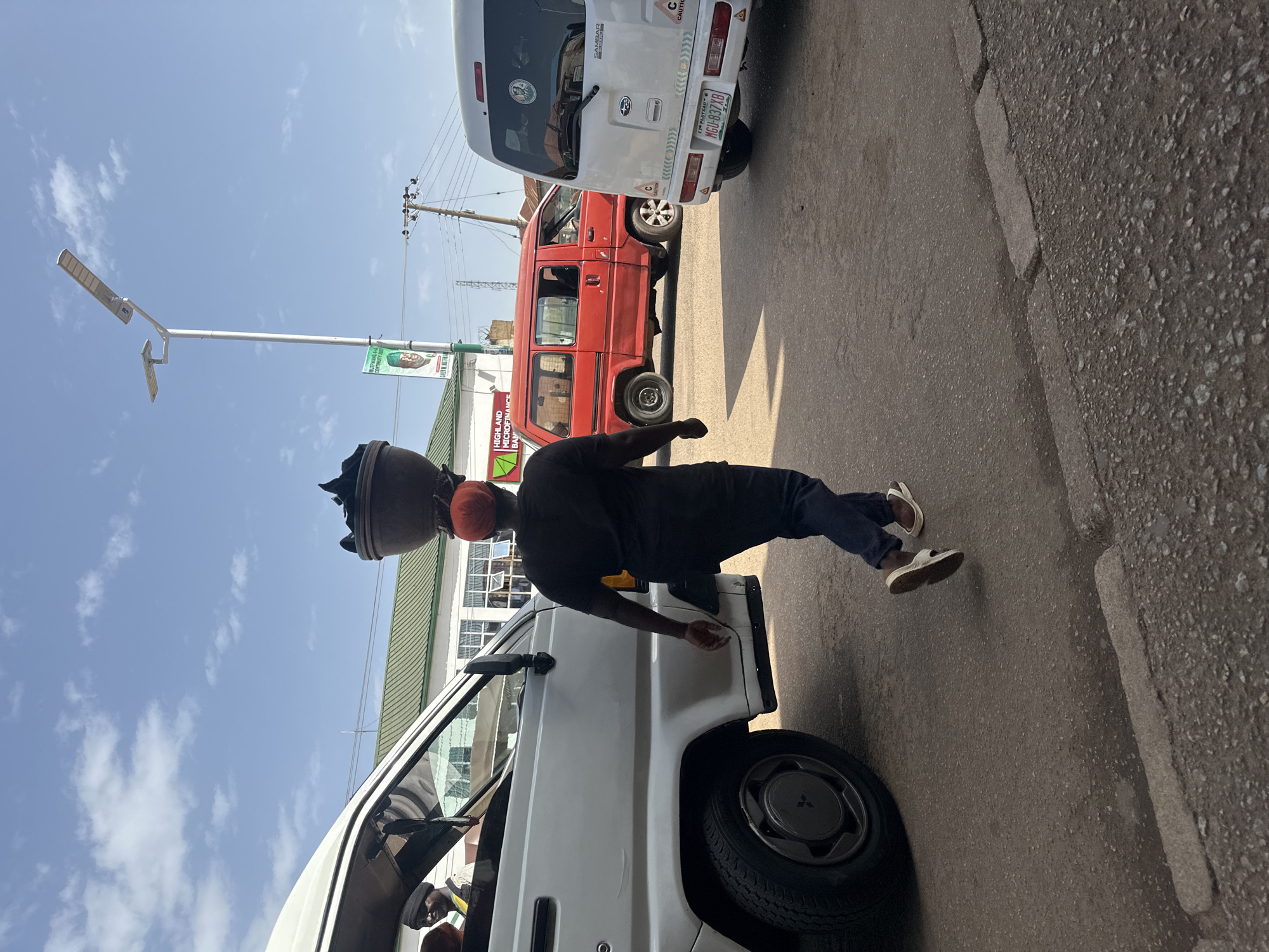 In Bukuru, Nigeria, a person wearing a black t-shirt, dark blue pants, and sandals walks away from the viewer, carrying a large, dark, bowl-like object on their head. The person is wearing an orange headscarf under the object. The scene is on a paved road, with a white van to the left and a red van and a white van to the right. The white van on the left appears to be a commercial vehicle, with a driver wearing a black headscarf visible through the window. In the background, a building with a green roof and "HIGHLAND MICROFINANCE BANK" signage is visible. A street lamp stands to the right of the building, and a poster featuring a man in a green shirt is attached to its pole. The sky is blue with scattered clouds.