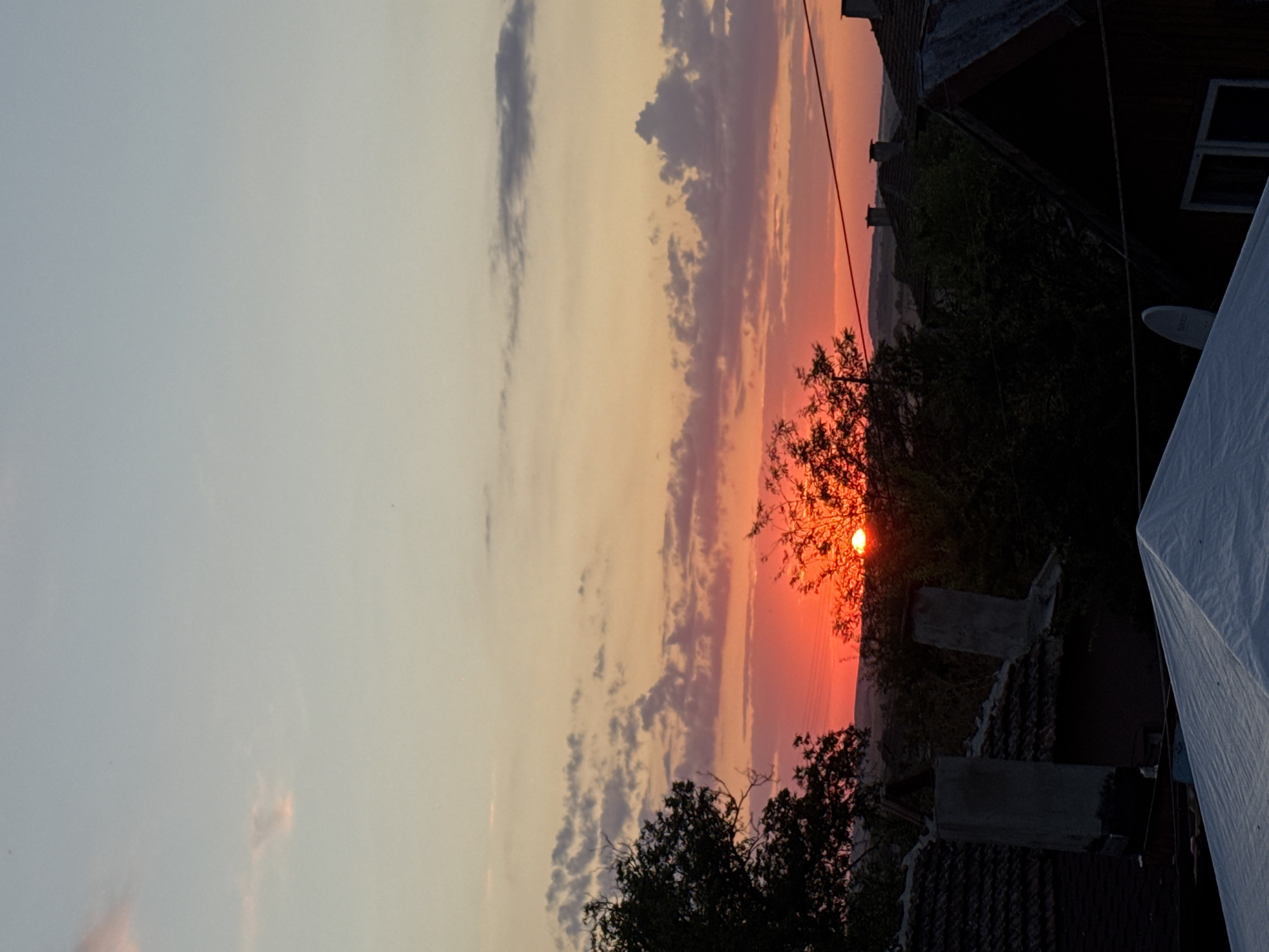 The image shows a sunset over Sredets, Bulgaria. The sun is setting behind trees and houses, casting a warm glow across the sky. The sky is filled with colorful clouds, ranging from pink and orange to purple and gray. The foreground is dominated by the rooftops of several houses, some with chimneys. Trees are visible on the left and right sides of the image, with some branches silhouetted against the sky. In the lower part of the image, a white tarp covers a structure. Power lines are also visible stretching across the scene. The overall impression is one of a peaceful evening in a rural setting.