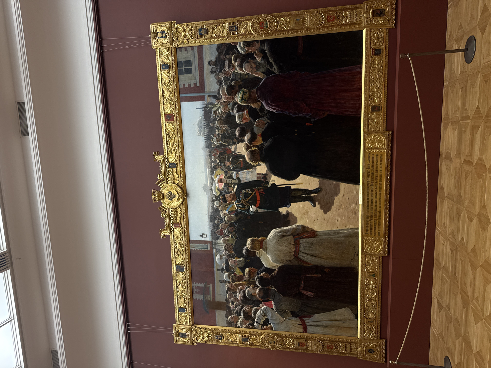 A large, rectangular, framed painting hangs on a burgundy wall in what appears to be a museum. The frame is elaborate and golden, featuring detailed carvings, coats of arms, and inscriptions. The painting depicts a crowd of people in what seems to be an outdoor event.
In the center of the crowd, an individual wearing a dark military uniform with gold accents is the apparent focus. The crowd includes men and women dressed in attire that is likely reflective of the era in which the painting was made.
The background suggests a city scene, with architectural elements that could potentially be Moscow. There is a building with red brick and white trim.
The room in which the painting hangs has a wooden parquet floor. A thin support line extends from the lower right corner of the frame towards a pole with a black disk at the floor. Wires suspending the frame are visible at the top corners.