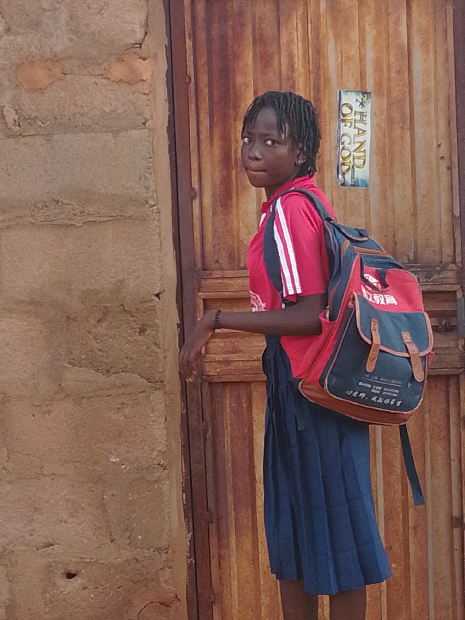 A young girl with braided hair is standing in front of a corrugated metal door in Gombe, Nigeria. She is wearing a red school uniform shirt with white stripes on the sleeves and a blue pleated skirt. She has a red and blue backpack with brown leather accents. The backpack has some writing on it in what appears to be Chinese characters. To the left of the girl is a rough, textured wall. A sign with "HAND OF GOD" written on it in English is attached to the corrugated door to the right of the girl.