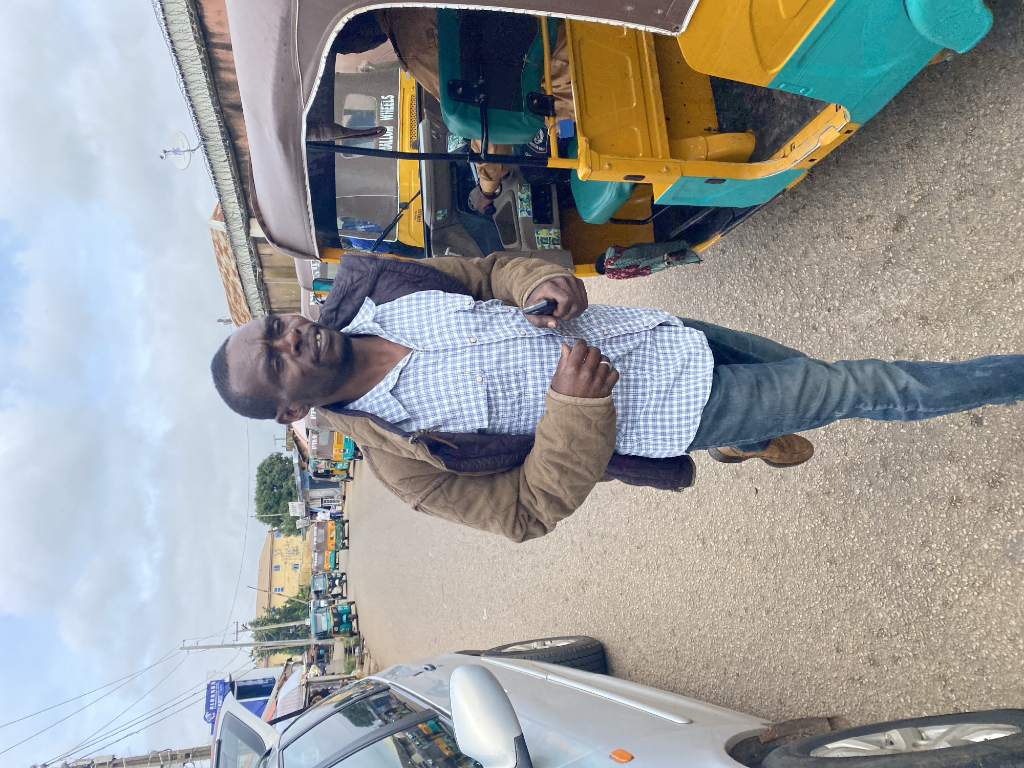 A man in a blue and white checkered shirt and brown jacket is walking down a street in Jos, Nigeria. He is holding a cell phone in his right hand. There are several auto-rickshaws, or Keke Marwa, parked on the side of the road. A silver car is parked on the left side of the road. The sky is cloudy.