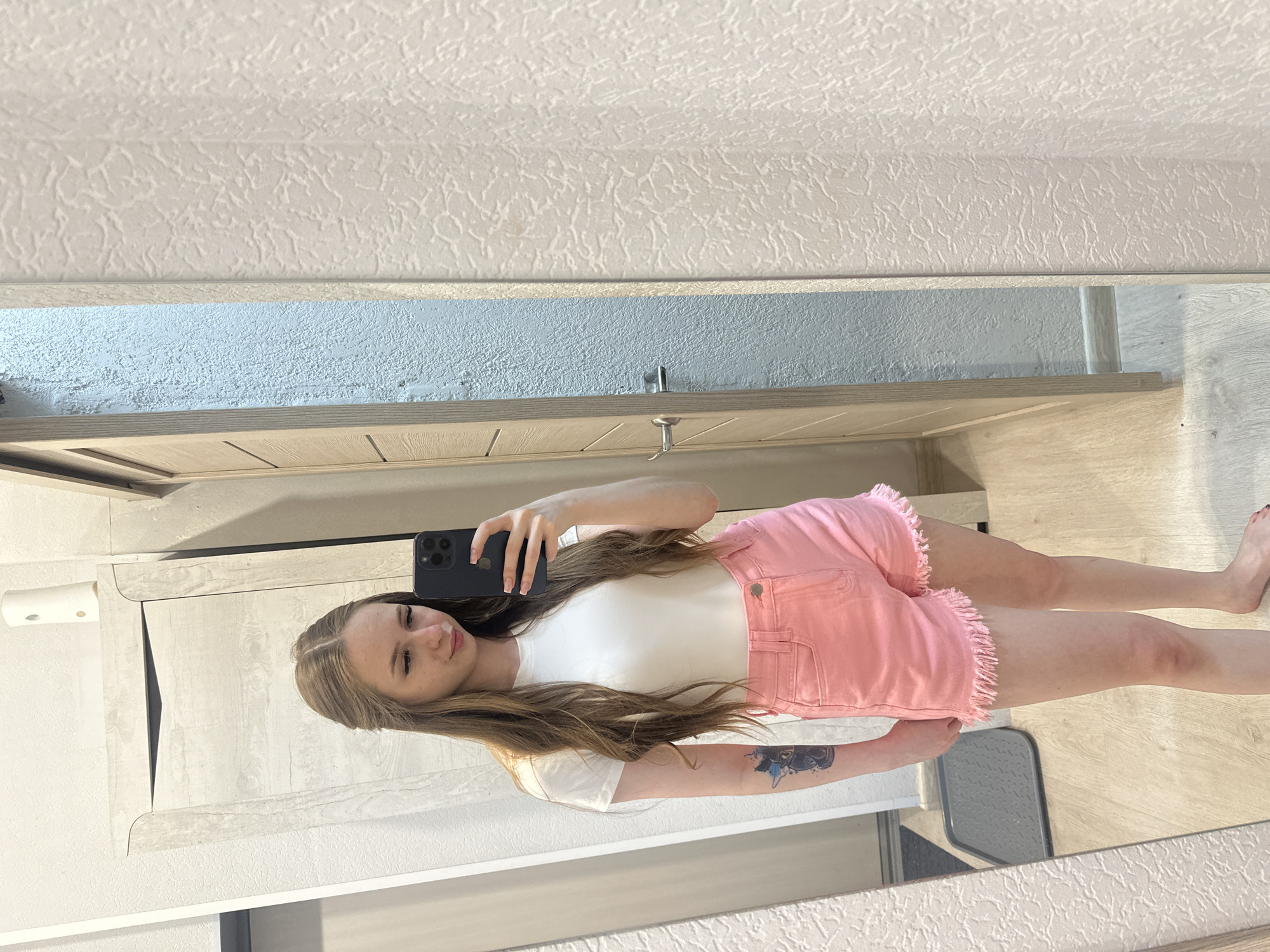 A young woman with long, wavy blonde hair, wearing a white t-shirt and pink denim shorts, takes a selfie in a mirror. She has a tattoo on her left arm. The background shows a wall, a door, and a mirror reflecting the room. The floor is wooden. The scene appears to be indoors, possibly in a residential setting.