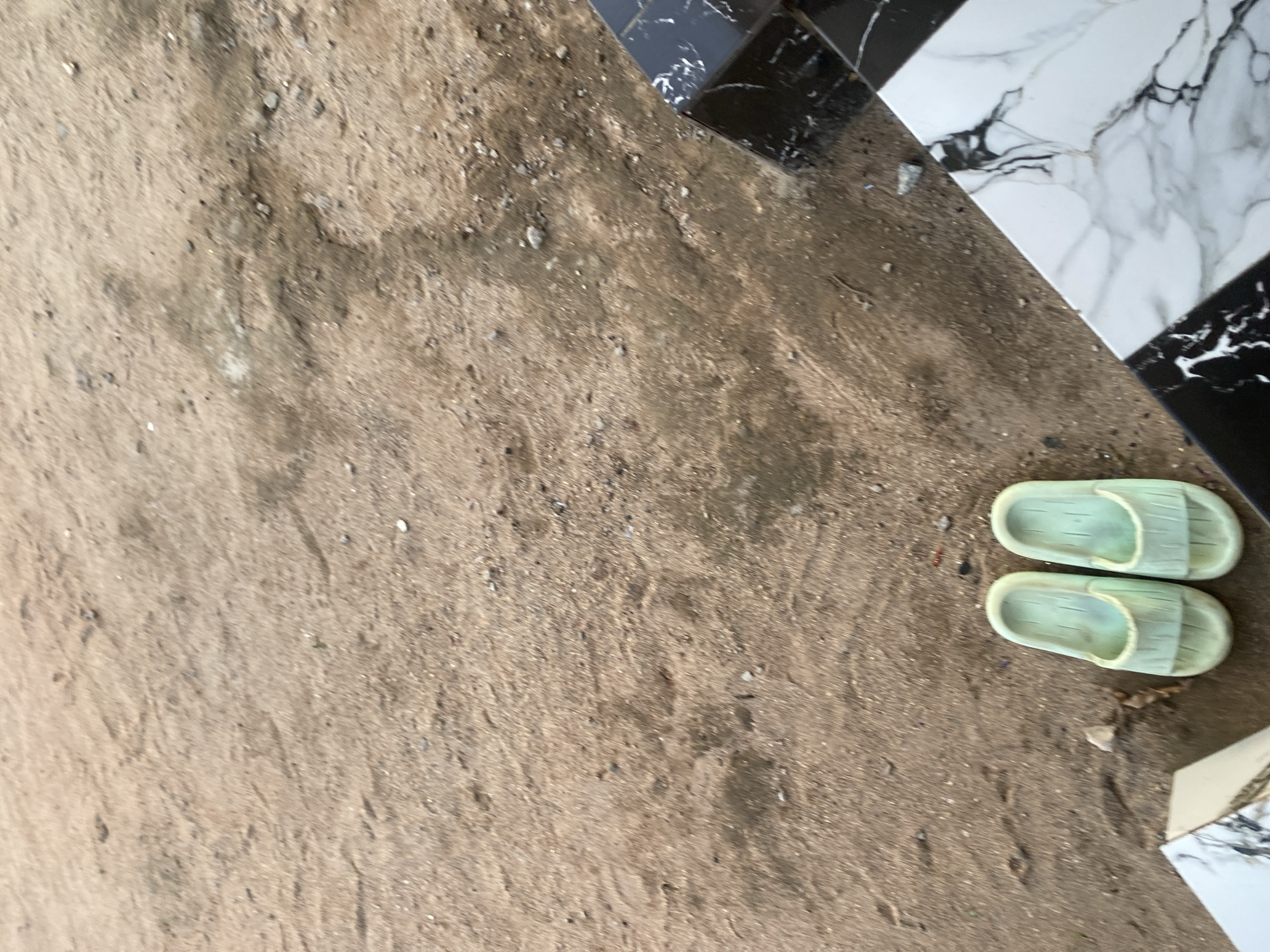 The image shows a pair of light green rubber slides resting on sandy ground. To the right, a surface with a black and white marble pattern is visible, suggesting a tiled area or a decorative object. The background consists of dry, sandy earth with small pebbles and debris scattered across it. The scene appears to be outdoors in a dry, possibly dusty environment. There is no indication of specific structures or landmarks that would pinpoint the location to Jauro Yinu, Nigeria, beyond what is stated.