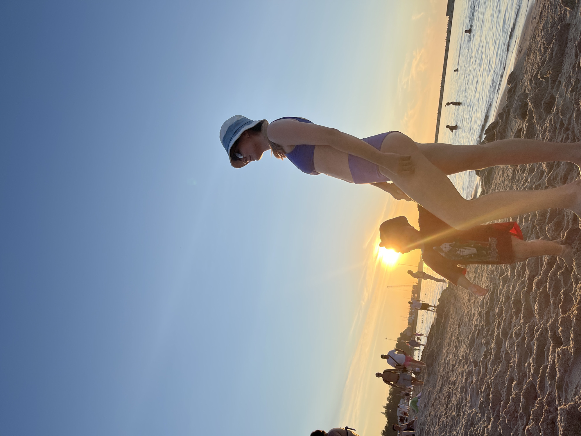 This is a vertical, low-angle, eye-level shot of a beach scene in Pionersky, Russia, during sunset. The sky is a gradient of clear blue near the viewer to a soft orange and yellow as it approaches the horizon, indicating it is late afternoon or early evening. The sun is visible as a bright, glowing orb low on the horizon, casting a warm light and creating lens flare. The sea is calm and reflects the sunset colors. The foreground is dominated by sandy beach with visible texture.
In the immediate foreground, a person wearing a blue and white bucket hat and a purple bikini is captured mid-action, seemingly leaning forward with their body parallel to the ground. Their face is in profile, looking down. Just behind and slightly to the right of this person, another individual is visible from the waist up, wearing a red t-shirt with a graphic print and a dark-colored hat or hood. Their face is obscured by the sun's glare.
Further back, other people are scattered across the beach and in the shallow water. Some are wading, while others appear to be walking or standing. In the distance, a long pier or breakwater extends into the sea, with a line of figures silhouetted against the bright sky at its edge. The overall mood is relaxed and summery, with a sense of people enjoying a pleasant evening at the seaside. There is no visible text in the image.