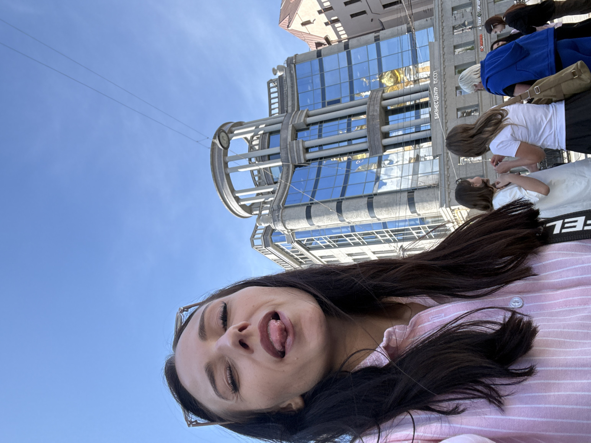 This media file captures a playful selfie taken by a young woman in an urban setting during the daytime. The image, rotated 90 degrees clockwise, shows a clear blue sky dominating the upper portion and a modern city building to the left, with several people visible in the background.
The main subject is a young woman, positioned prominently on the right side of the frame (when viewed correctly), looking downwards and slightly to the left. She has long, dark hair and is wearing a pink and white striped button-up shirt. Her expression is lighthearted and silly, with her eyes mostly closed and her tongue sticking out. What appears to be the arm of eyeglasses is visible near her temple.
In the background, on the left side, stands a large, multi-story building featuring a contemporary design with extensive blue-tinted glass windows that reflect the bright sky and possibly other structures. The building's facade is adorned with distinctive architectural elements, including large, curved, metallic-colored structures resembling pipes or framing, possibly part of its support or decorative features. Visible text on the building, in Cyrillic, reads "БИЗНЕС ЦЕНТР" (BUSINESS CENTER) and "ЦСОП."
Below the building and towards the center, several other individuals are visible on what appears to be a street or plaza. They are engaged in typical urban activities, either walking or standing. One person has blonde hair, and another is wearing a distinct blue jacket and carrying a brown shoulder bag.
The scene is bathed in bright, natural light, indicating a sunny day. The sky is largely cloudless, suggesting clear weather. Two thin wires are faintly visible stretching across the blue sky. The presence of Cyrillic text on the building confirms the user-provided context of Saint Petersburg, Russia. The overall mood conveyed by the main subject is one of casual fun.