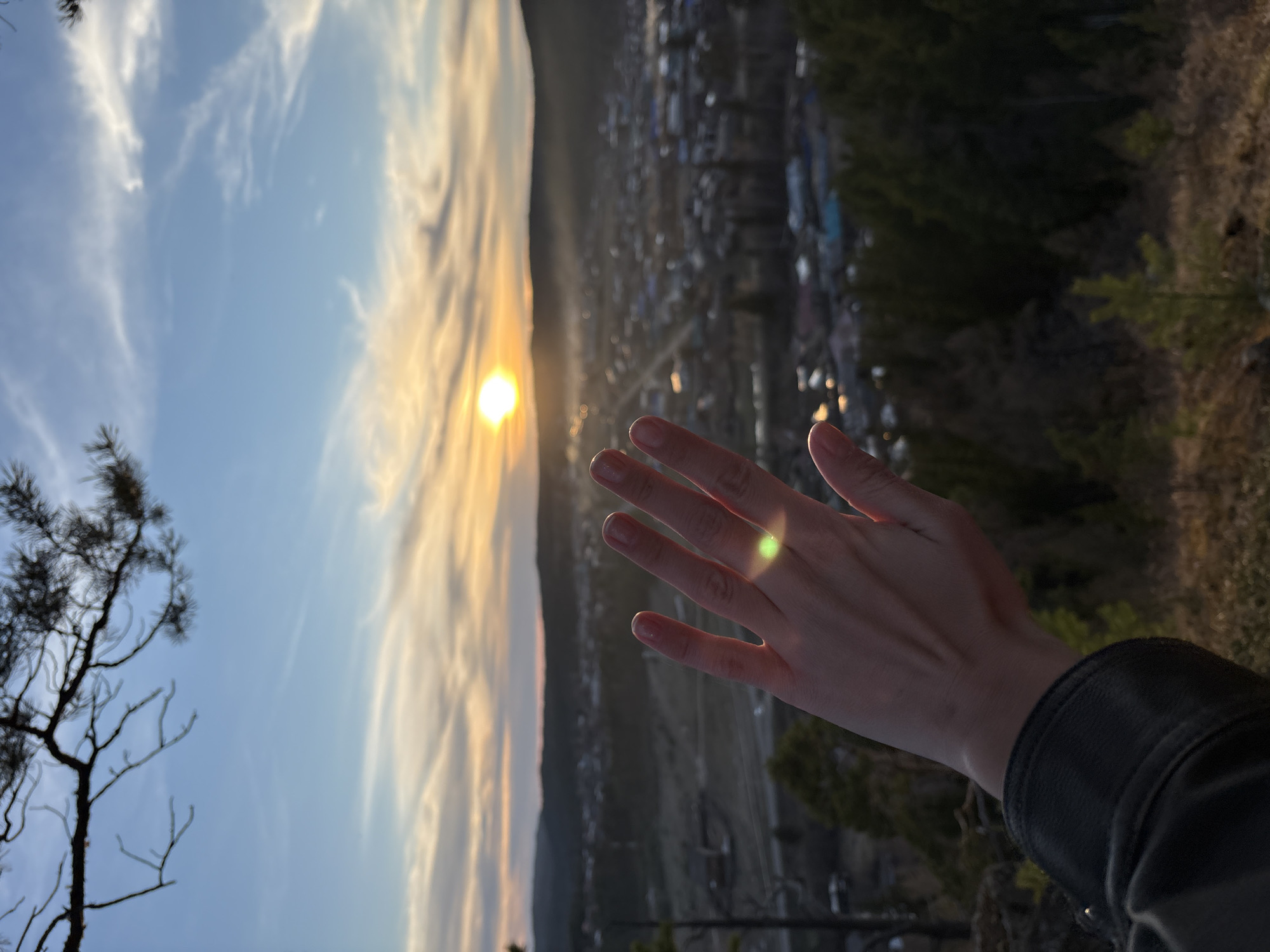 A human hand reaches out toward a low sun partially obscured by clouds in Невон, Russia. The background shows a town and forest.