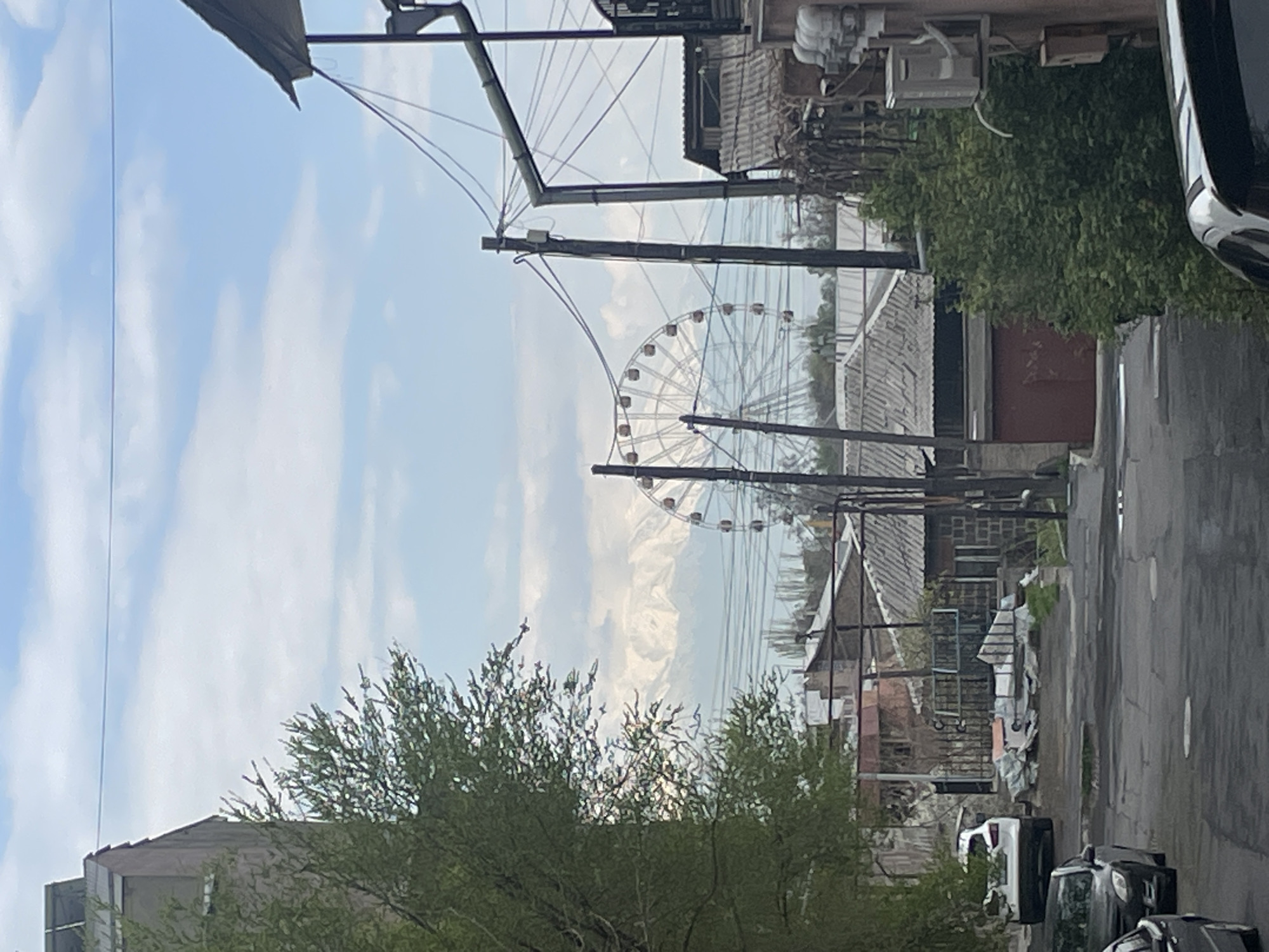 The image depicts a street in Yerevan, Armenia, with a Ferris wheel visible in the distance between buildings and utility poles. The street appears narrow and residential, with parked cars on the right and buildings on both sides. Overhead, power lines crisscross. Vegetation, including trees, is visible along the street. The sky is light blue with scattered clouds. The image is slightly tilted.