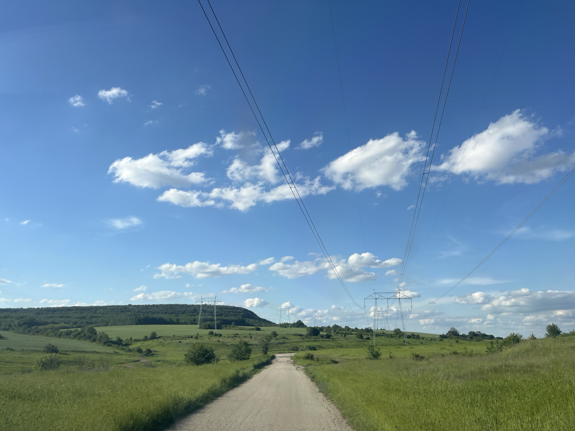 A ground-level shot showcases a dirt road stretching into the distance, flanked by green fields. Power lines run overhead, supported by several transmission towers visible along the horizon. A wooded hill forms a backdrop against a bright blue sky dotted with scattered clouds. The location is unknown.