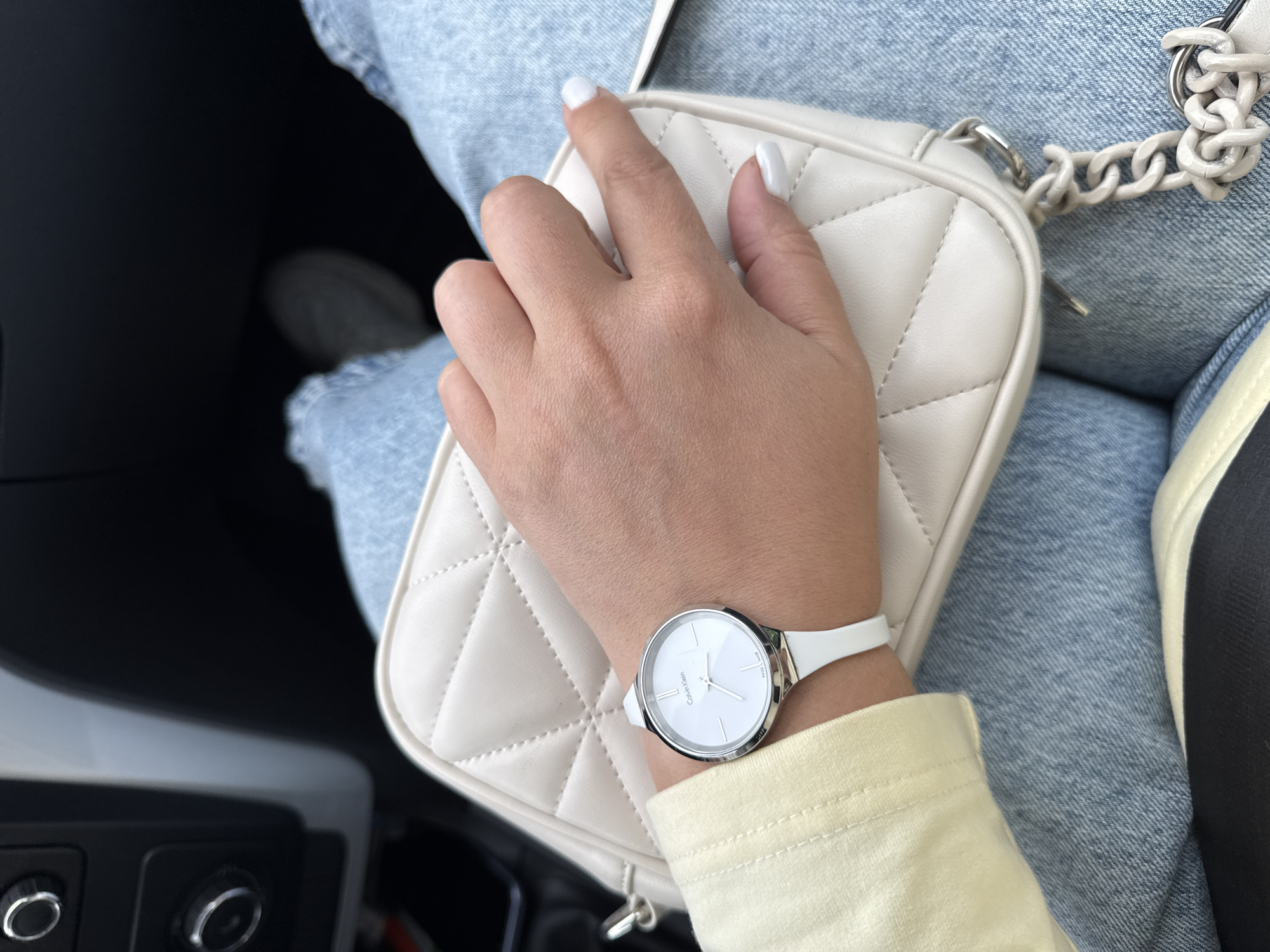 A close-up shot captures a woman's left hand resting on a cream-colored quilted purse. She is wearing a white analog watch with a silver bezel and a white strap, positioned on her wrist. The watch face is white with silver hour markers and hands, and the brand name "Calvin Klein" is visible. She has white nail polish on her fingernails. The woman is wearing light blue denim jeans and a light yellow long-sleeved shirt. The purse has a quilted diamond pattern and a chunky white chain strap. In the background, a dark interior, possibly of a car, is visible, with some controls and a seat. The scene appears to be indoors. There is no indication of time of day, weather, or specific location beyond the general interior setting. No emotions are conveyed.