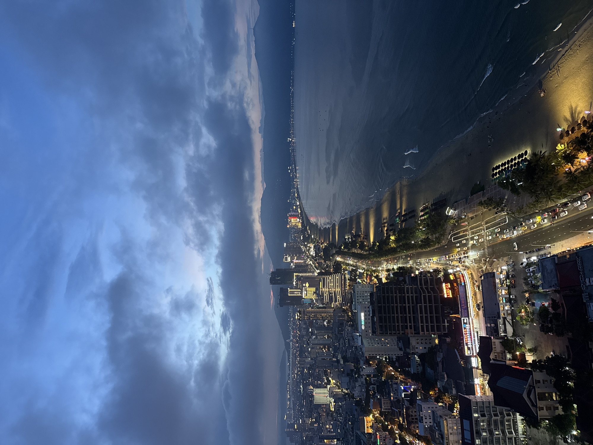 This is a high-angle, wide shot of the coastline of Đà Nẵng, Vietnam, taken at dusk or early evening. The sky is a deep blue with dramatic, heavy clouds. On the left, a dense urban cityscape stretches into the distance, filled with illuminated buildings of varying heights, including prominent high-rises. A main road runs along the coastline, lined with streetlights, palm trees, and a steady flow of vehicle traffic, indicated by streaks of light. On the right, a wide sandy beach is visible, with rows of umbrellas and loungers, some lit by artificial lights. The ocean meets the beach, with gentle waves breaking along the shore. The overall scene depicts a vibrant coastal city at twilight, with the lights of the city reflecting on the water.