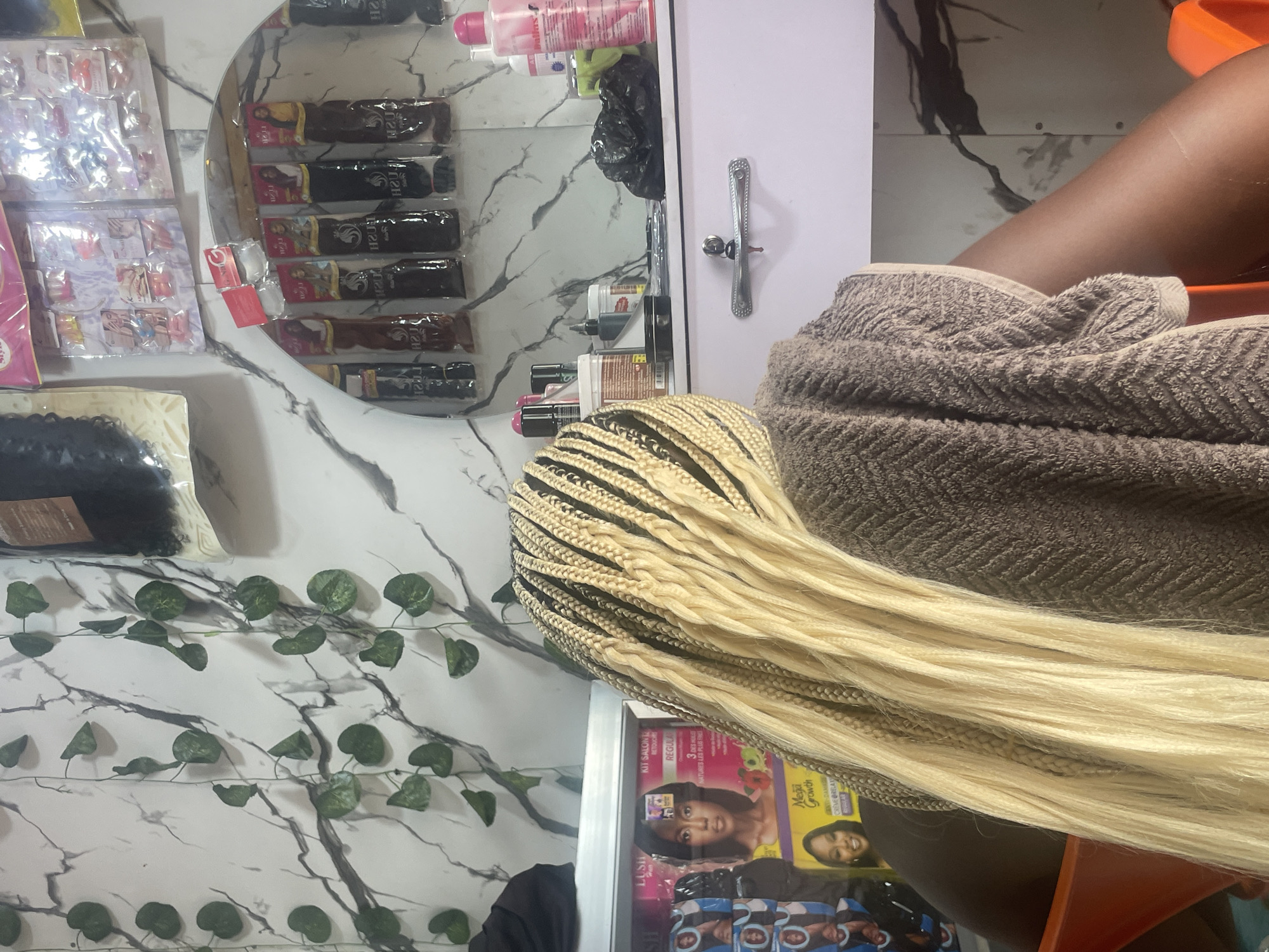 In a hair salon located in Lafia, Nigeria, a person with long, blonde braids is seated, facing away from the camera and towards a vanity. The braids, which appear to be synthetic extensions, are intricately woven and cascade down the person's back. A brown towel is draped over the person's shoulders, covering their upper back. The vanity behind the person is cluttered with various hair care products, including bottles of lotion and sprays. A mirror above the vanity reflects shelves stocked with packaged hair extensions in different colors and styles. The walls of the salon are decorated with a marble-patterned wallpaper and artificial ivy vines, adding a touch of greenery to the space. The overall impression is one of a busy salon environment focused on hair styling and maintenance.