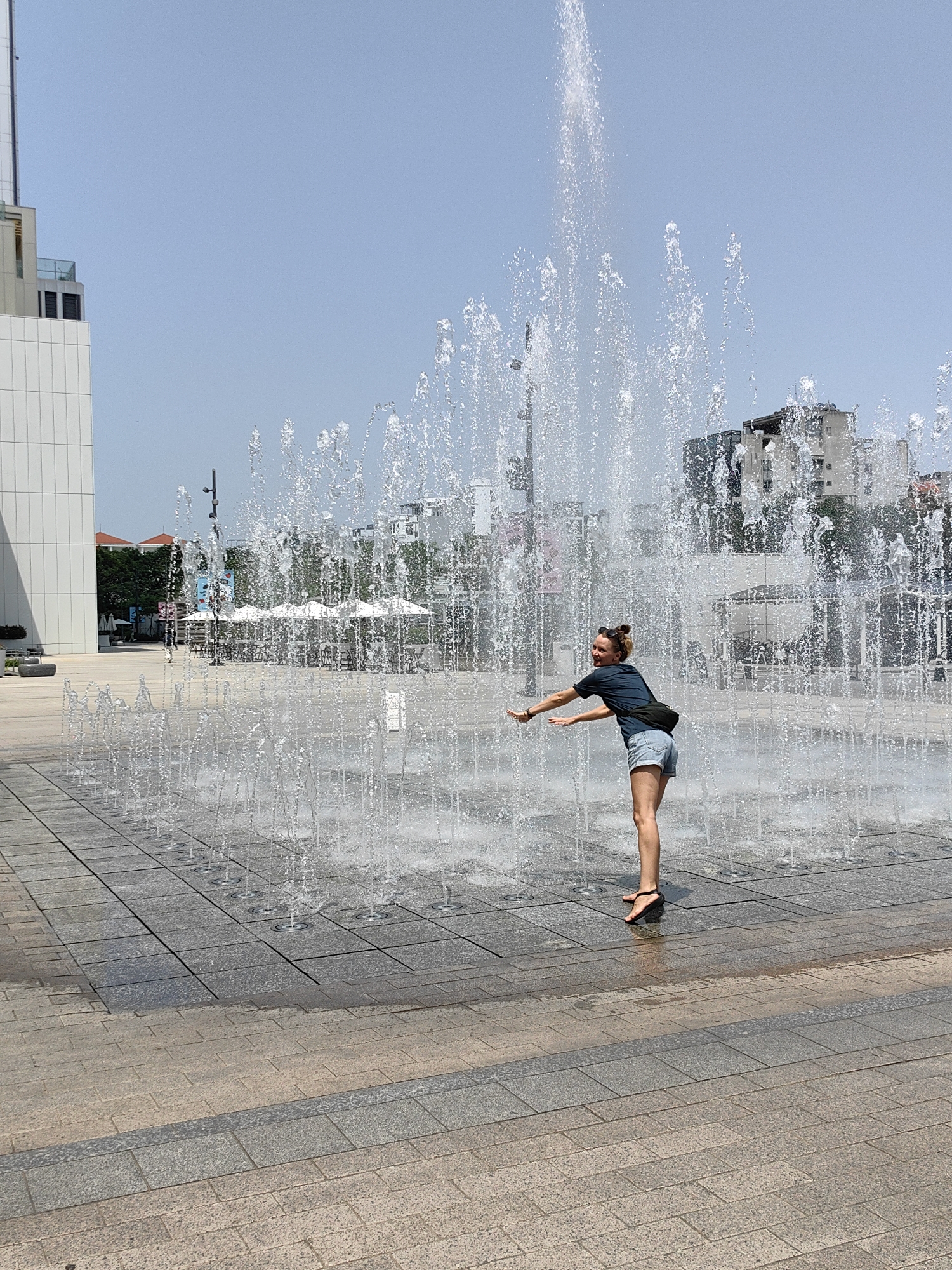A young woman is captured mid-action, playfully interacting with a ground-level water fountain in a public square in Hà Nội, Vietnam. She has light skin, her hair tied in a bun, and is dressed in a dark t-shirt, light denim shorts, and black sandals, with a black fanny pack worn across her body. Her arms are outstretched, and she leans slightly, looking back over her right shoulder with a happy, smiling expression, seemingly enjoying the water spray from the numerous jets.
The fountain consists of many individual streams of water shooting upwards from the paved ground, creating a dynamic backdrop around the woman. The plaza is extensively paved with light and dark gray rectangular and square tiles, with water splashes visible on the surface. In the background to the left, a modern white building with geometric lines and some glass elements rises under a clear, bright blue sky. To the right, a cityscape of older, multi-story buildings with various hues and some red roofs is visible. A cafe area with white umbrellas, tables, and chairs can be seen in the mid-ground to the left, partially obscured by the fountain. The overall scene depicts a cheerful and refreshing moment on a sunny, clear day.