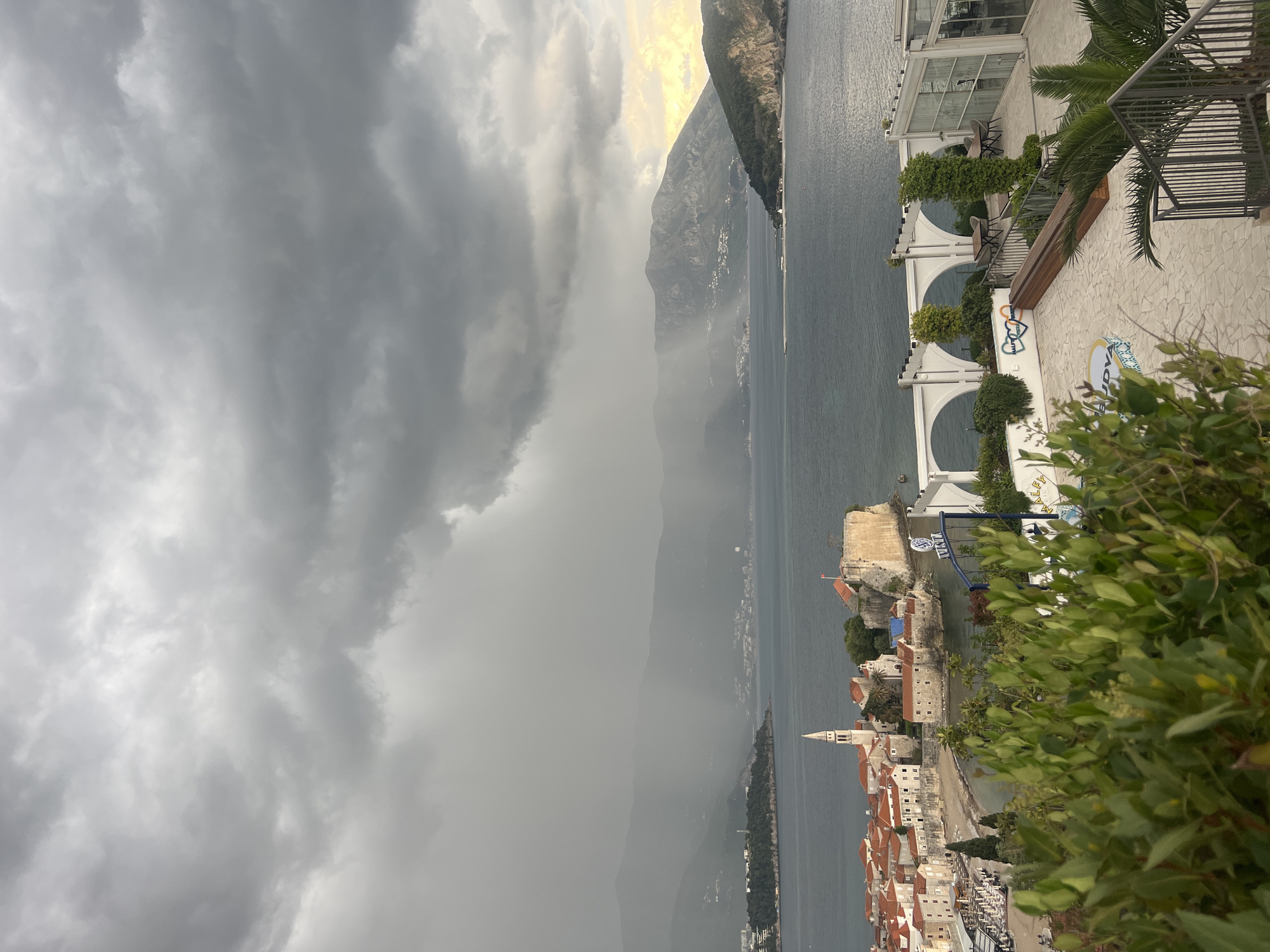 This media file presents a panoramic view of a coastal town, widely recognized as Budva, Montenegro, nestled against the base of a mountain range and bordering a large body of water. The scene is dominated by a dramatic, heavily overcast sky with dark, stormy grey clouds. Rain or mist appears to be falling over the distant mountains on the left side, obscuring them significantly. A lighter, almost golden hue can be seen through a break in the clouds over the mountains on the right, hinting at either approaching sunlight or the storm's edge.
The water, likely the Adriatic Sea, is a dark, expansive body, reflecting the somber sky. The historical town of Budva, located in the mid-ground, features dense clusters of buildings with characteristic reddish-orange tiled roofs. A tall, slender white bell tower with a pointed spire stands prominently within the town's architecture, and old stone fortifications line parts of the coastline.
In the foreground on the right, a modern, multi-tiered building, likely a restaurant or hotel, showcases white arched structures, glass enclosures, and outdoor terraces furnished with tables and chairs. Lush green vegetation, including palm trees and various bushes, frames this foreground area. A decorative paved area is also visible. On a wall of this modern building, a mural-like design includes the text "BUDVA" along with stylized heart shapes and other artistic elements.
The overall mood is dramatic and powerful, with the stormy weather creating a striking contrast between the dark sky, the grey mountains, and the warm-toned roofs of the historic town. The time of day is ambiguous due to the heavy cloud cover, but the faint light suggests either dawn or dusk battling through the storm.