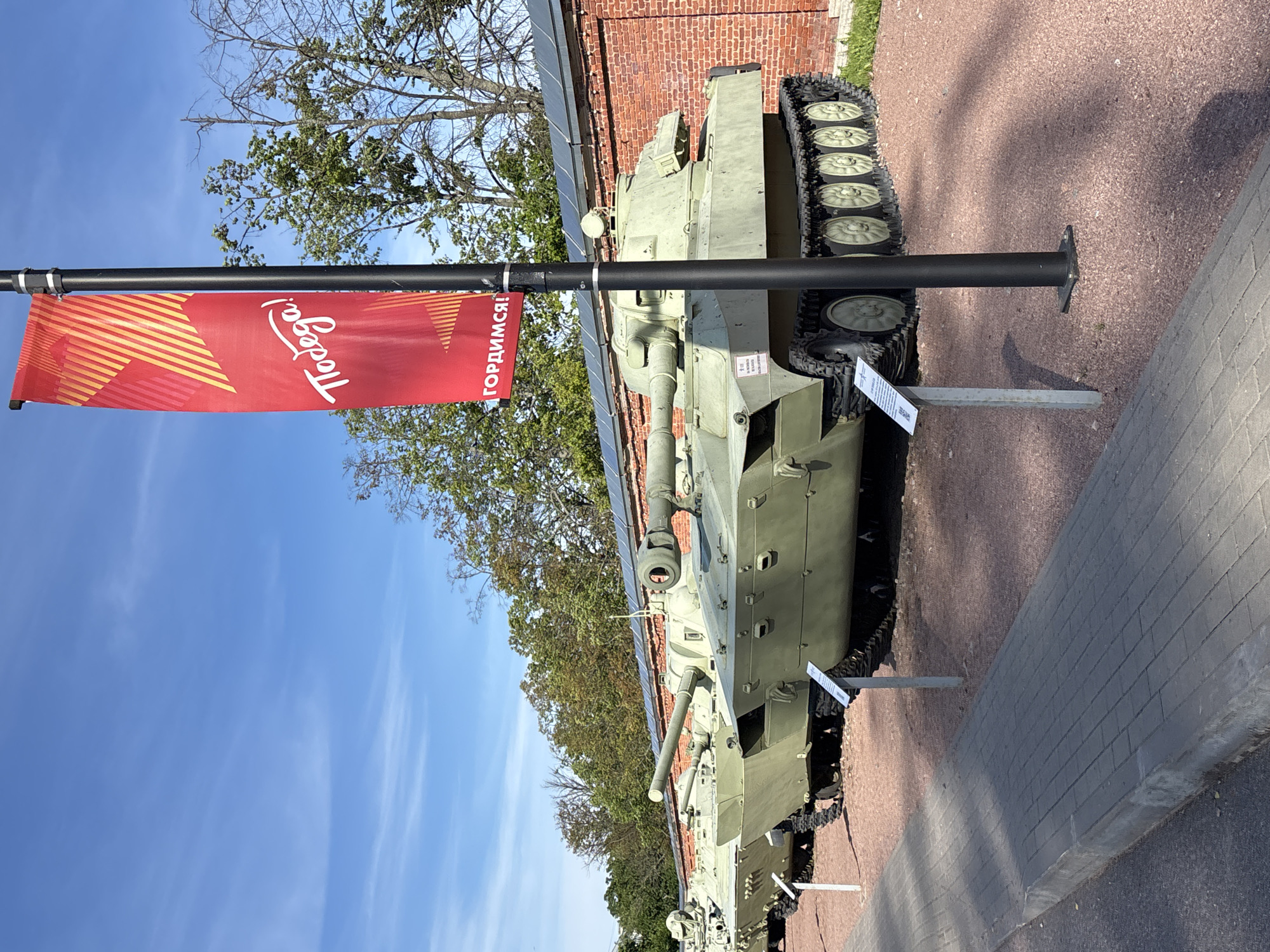 This image depicts a historical military vehicle, likely an armored personnel carrier, on display outdoors. The vehicle is painted in a military green camouflage and features prominent tracks. It is positioned on a paved walkway next to a gravel path. In the background, a red brick building and lush green trees are visible, suggesting a museum or historical site.
A red banner with white lettering is attached to a black pole in the foreground. The text on the banner reads "TODAY!" and "Гордимся!" which translates to "We are proud!". The banner has a graphic design of stylized diagonal lines in yellow and red.
The sky is a clear blue with scattered white clouds, indicating a pleasant day. The sunlight casts shadows on the ground, suggesting it is likely daytime, possibly late morning or early afternoon. The overall scene conveys a sense of historical preservation and perhaps public education about military heritage. The location context of Kronstadt, Russia, reinforces the likelihood of this being a military-related exhibit. There are no people visible in the image.