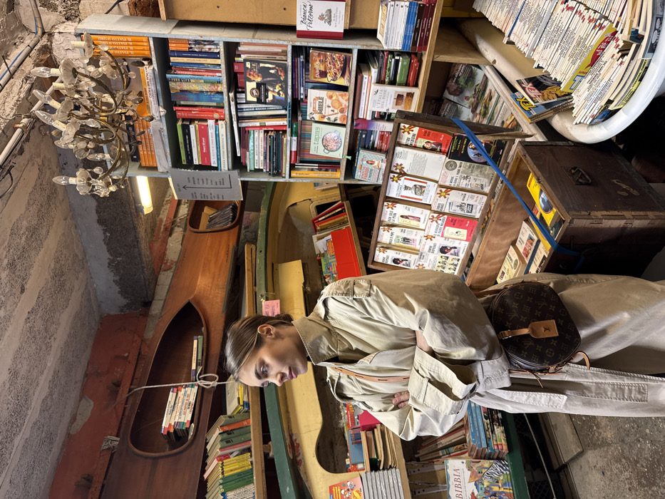 This image depicts the interior of Libreria Acqua Alta, a unique bookstore in Venice, Italy. A young adult female, approximately in her late 20s to early 30s, stands in the foreground, slightly to the left of center. She is wearing a beige trench coat, buttoned, with her arms crossed in front of her. A circular Louis Vuitton monogrammed crossbody bag with a brown leather strap and luggage tag is slung across her body, resting at her right hip. Her head is turned to her left, facing away from the viewer, with a contemplative or observant expression. Her hair is pulled back from her face.
The bookstore environment is characterized by its cluttered and unconventional display of books. On the left, the halves of wooden boats are integrated into the wall as shelving, holding stacks of books. A white rope hangs from the ceiling near these boat structures. The ceiling itself is uneven, featuring exposed concrete and patches of reddish-brown paint, with a modern fluorescent light fixture and an antique-style multi-arm crystal chandelier. Numerous bookshelves, densely packed with books of various sizes, colors, and languages, line the walls. A tall, light-blue painted bookshelf occupies the right side of the frame, extending almost to the ceiling.
In the mid-ground, a wooden display rack holds rows of small paperback books, presented cover-out. Below this rack, an open wooden chest also contains books, secured with blue straps. To the far right, partially visible, is a white bathtub filled with stacks of books. A white sign with black text hangs from the rightmost bookshelf. The floor appears to be a dark, worn concrete or stone surface.