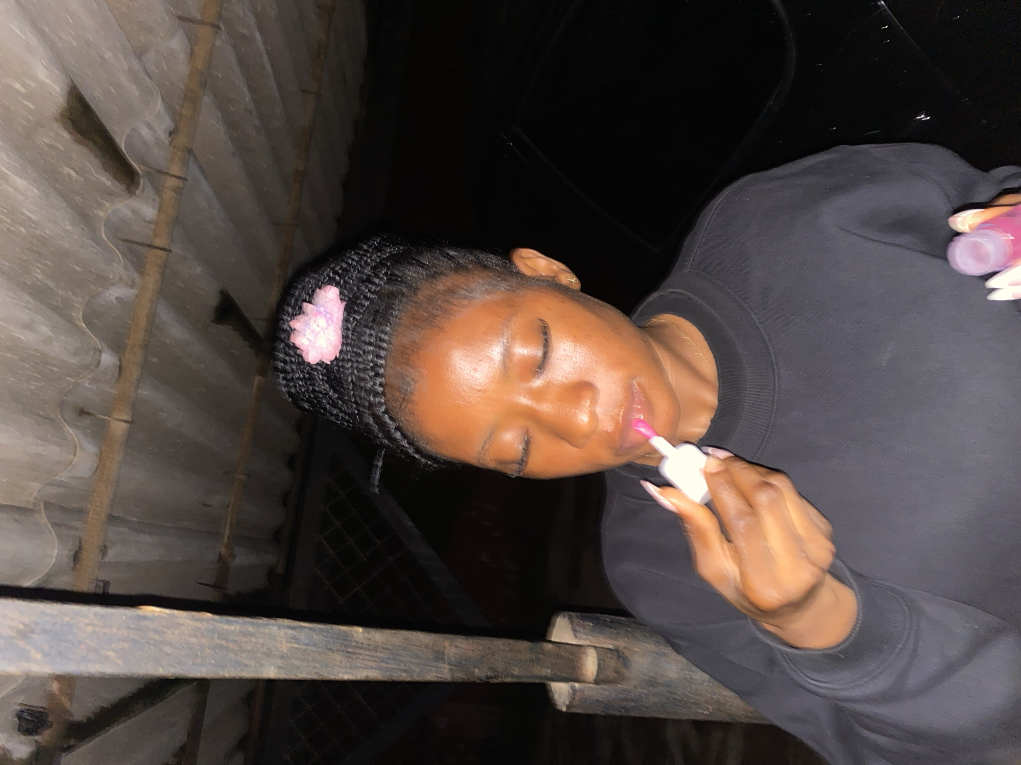 An African woman with braided hair and a pink flower accessory is applying lip gloss. She is wearing a black sweatshirt. The background shows a corrugated metal roof and a dark car. The scene appears to be outdoors at night, possibly in Jos, Nigeria.