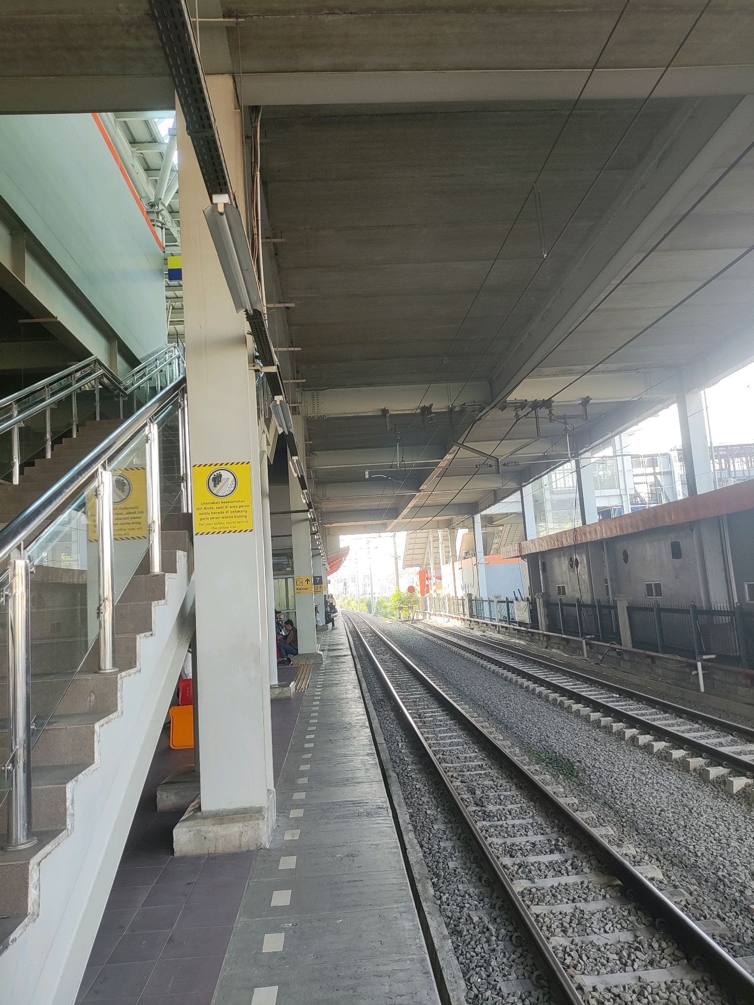 This is a daytime, eye-level, vertical shot of an outdoor train station platform in Bekasi, Indonesia. The platform is empty except for a few seated individuals in the background and a few scattered objects. The platform is made of grey tiles with a white dotted line marking the edge. To the left, there is a set of stairs with metal railings and glass panels. On the stairs, there are two yellow signs with black text and symbols, warning people to stay behind the yellow line for their safety. In the background, there are train tracks with ballast. The area is covered by a concrete roof supported by pillars. The lighting suggests it is either morning or afternoon, with sunlight filtering through the gaps in the roof.