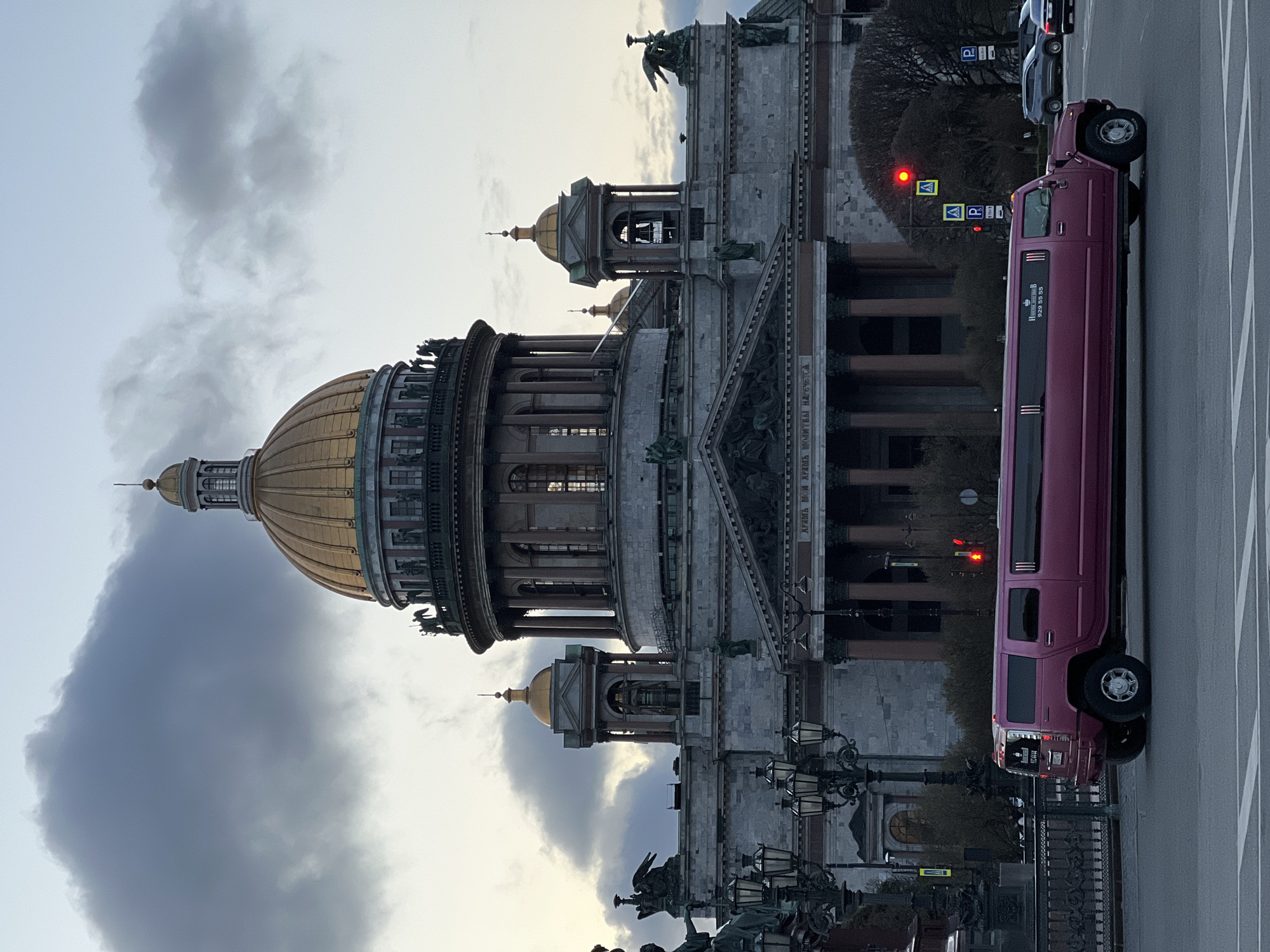 The image shows Saint Isaac's Cathedral in Saint Petersburg, Russia. The cathedral's large golden dome is visible. A long, pink limousine is parked on the street next to the cathedral. The sky is overcast with grey clouds. Street lamps and a section of a wrought iron fence are visible in the foreground. Several other cars can be seen on the street. There are no people visible.